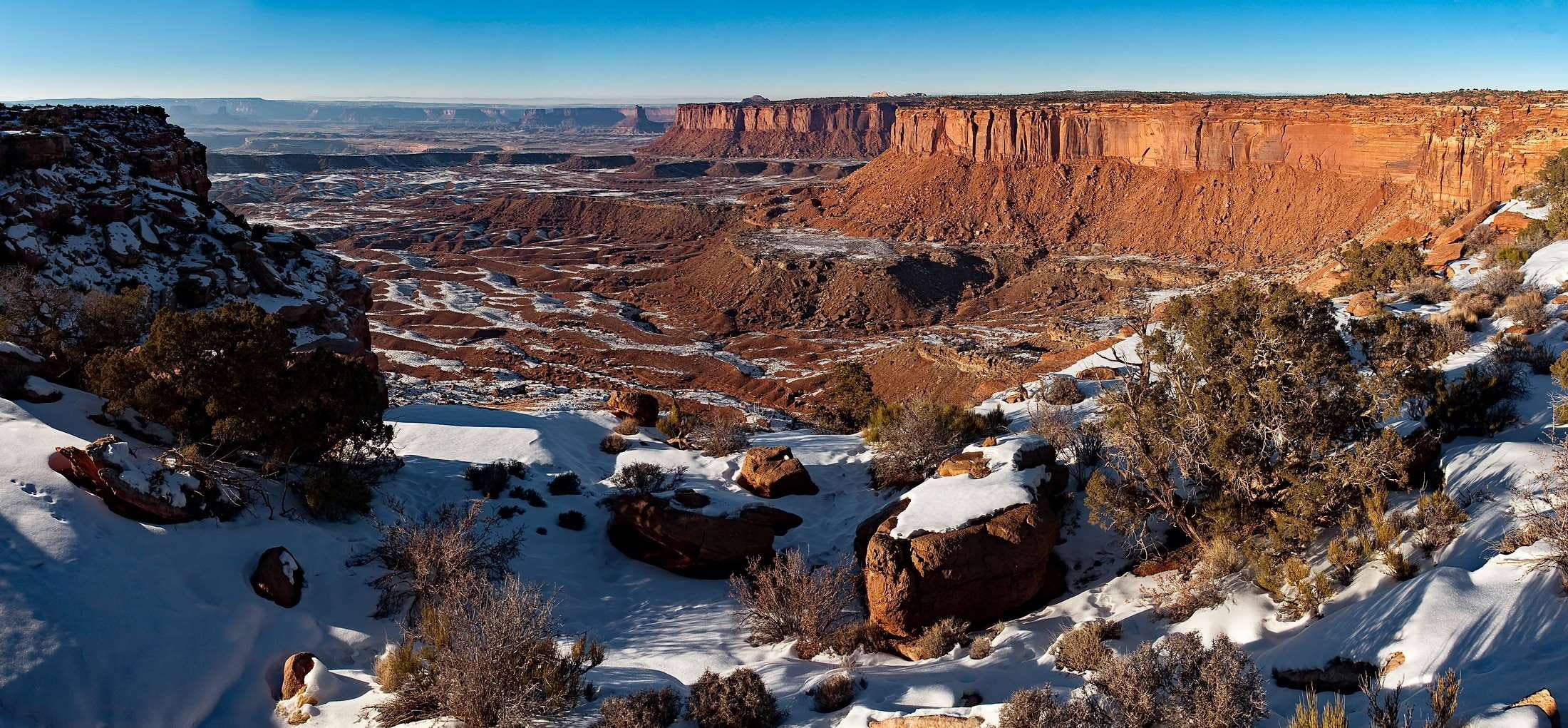 Canyonlands National Park 桌布 壁紙 — 在廣闊峽谷的清澈藍天下，白雪覆蓋了崎嶇的紅色岩層和稀疏的沙漠灌木。