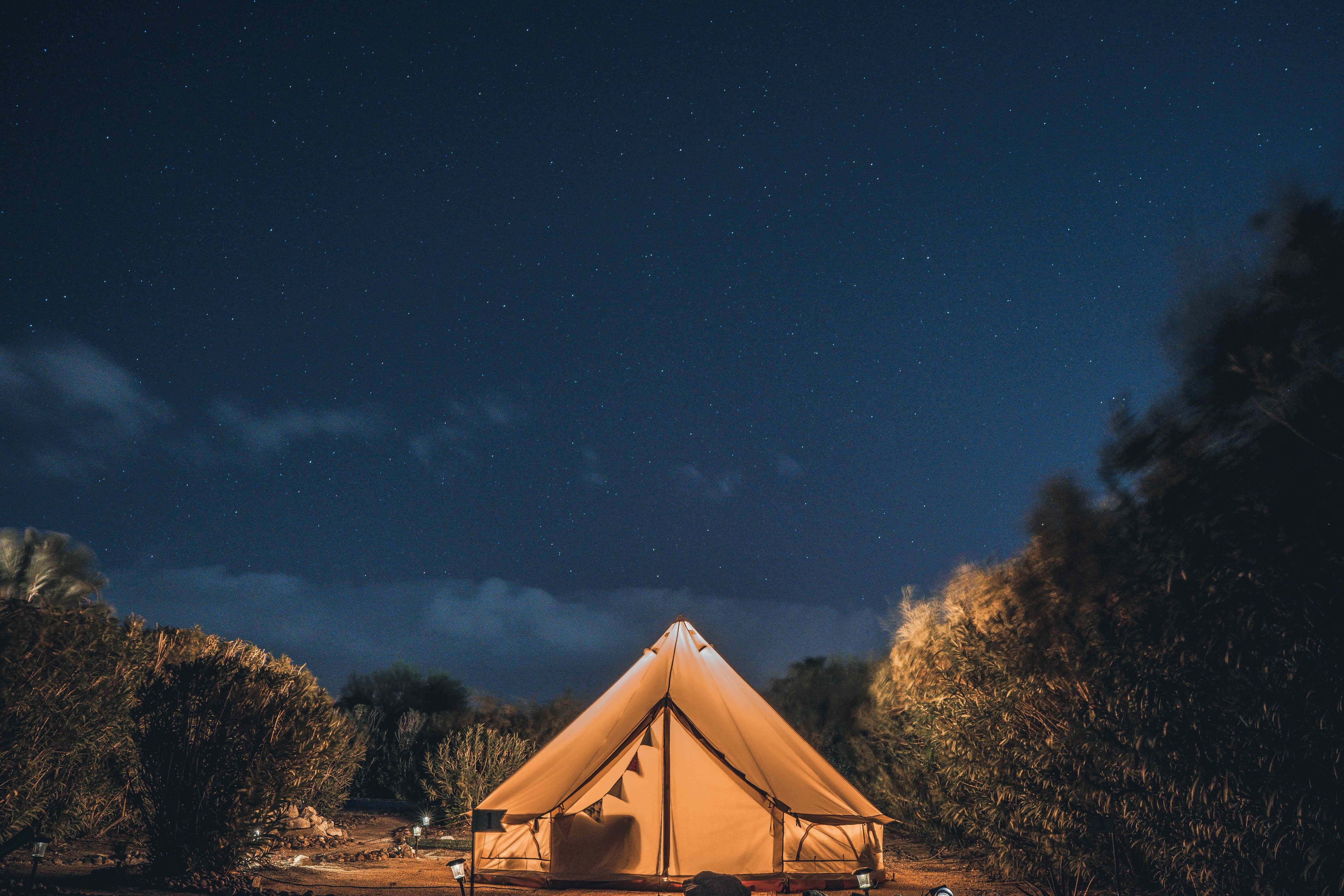 Camping 桌布 壁紙 — 在廣闊的星空下，一頂溫暖照明的鐘形帳篷發光，周圍是黑暗的樹木剪影。