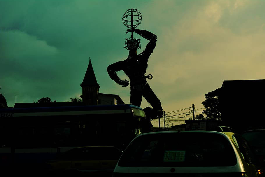 Silhouette 桌布 壁紙 — 在憂鬱的藍綠色和黃色天空下，一座高聳的雕像手持地球儀，矗立於有汽車和教堂尖頂的城市街道之上。