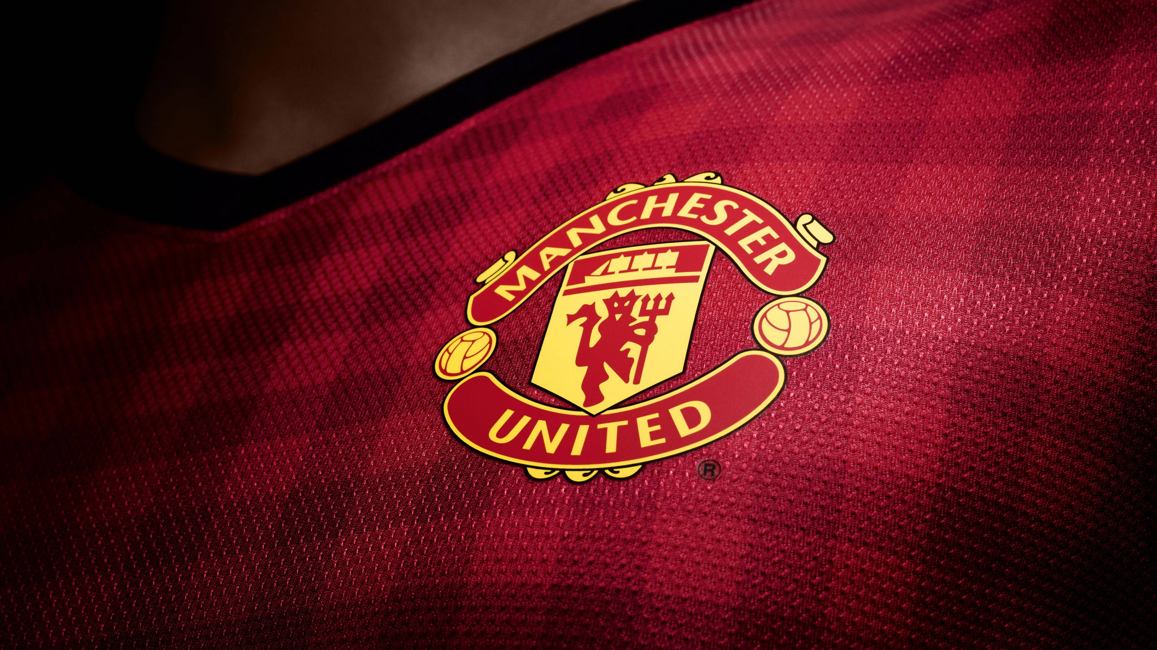 Manchester United Logo 桌布 壁紙 — 在戲劇性燈光下，曼聯會徽在帶有紋理的紅色足球球衣上的特寫。