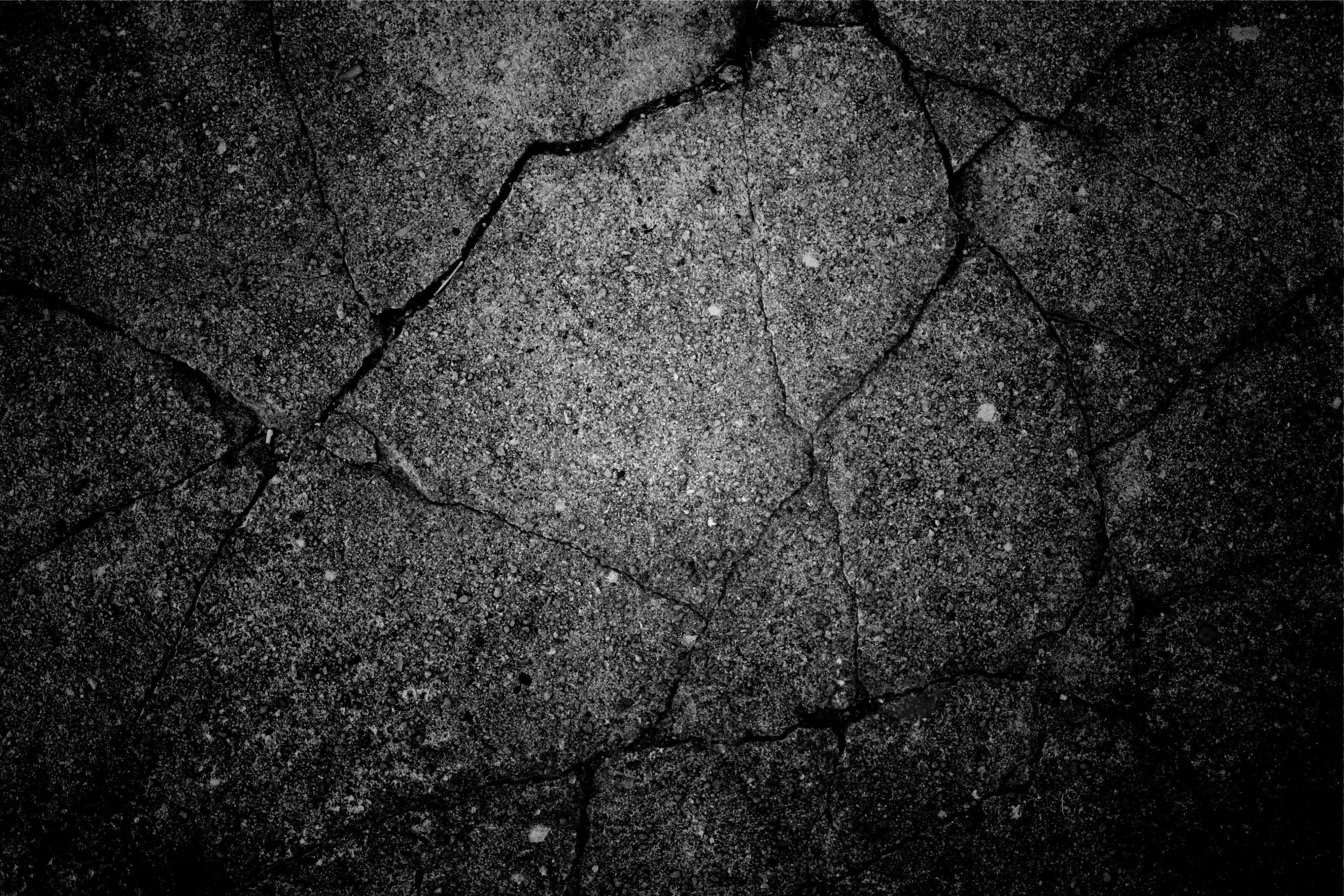 Dark Grunge 桌布 壁紙 — 在戲劇性燈光下，粗糙的單色特寫鏡頭，呈現出裂縫深且散佈斑點的柏油路面。