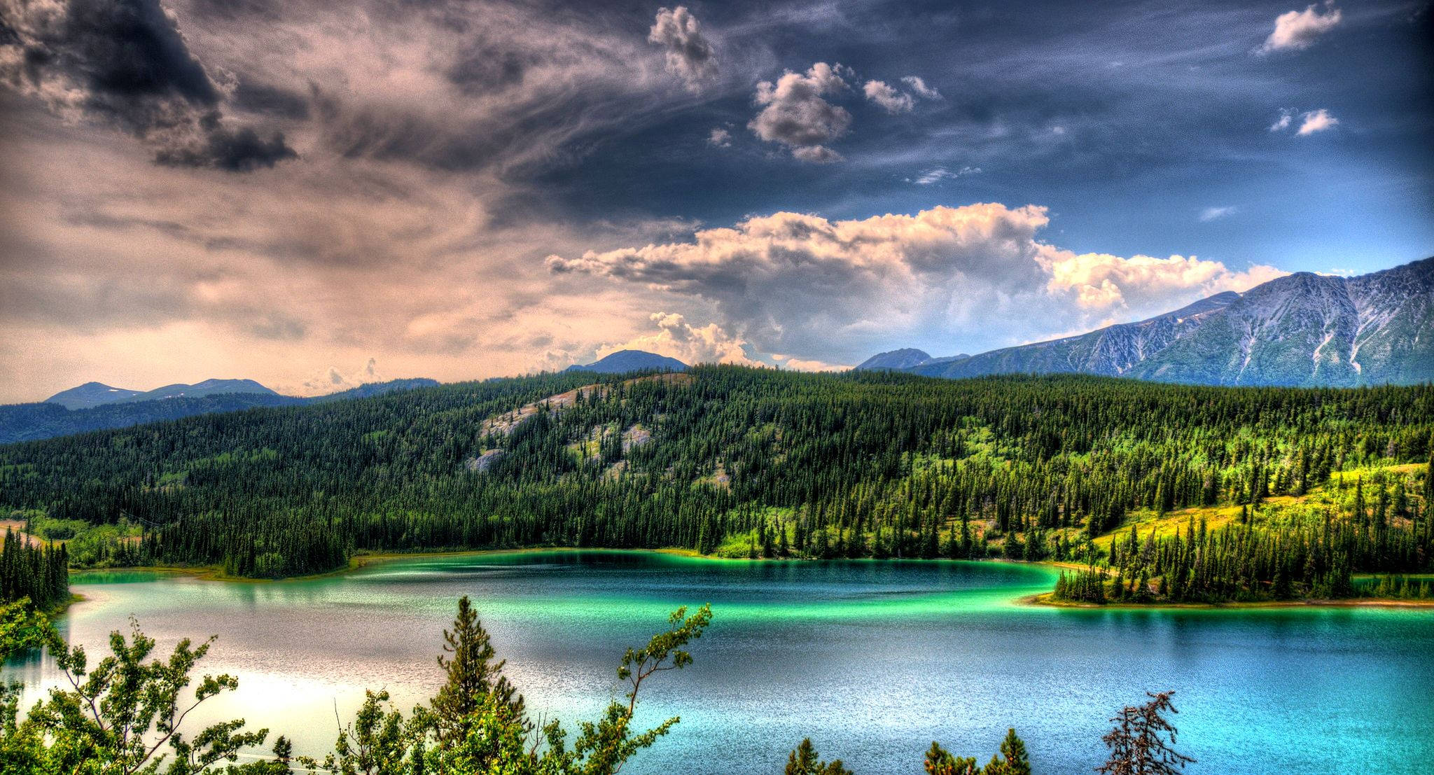 Lake 桌布 壁紙 — 在戲劇性的多雲天空下，一個充滿活力的藍綠色湖泊坐落在茂密的綠色森林和巍峨的山脈之間。