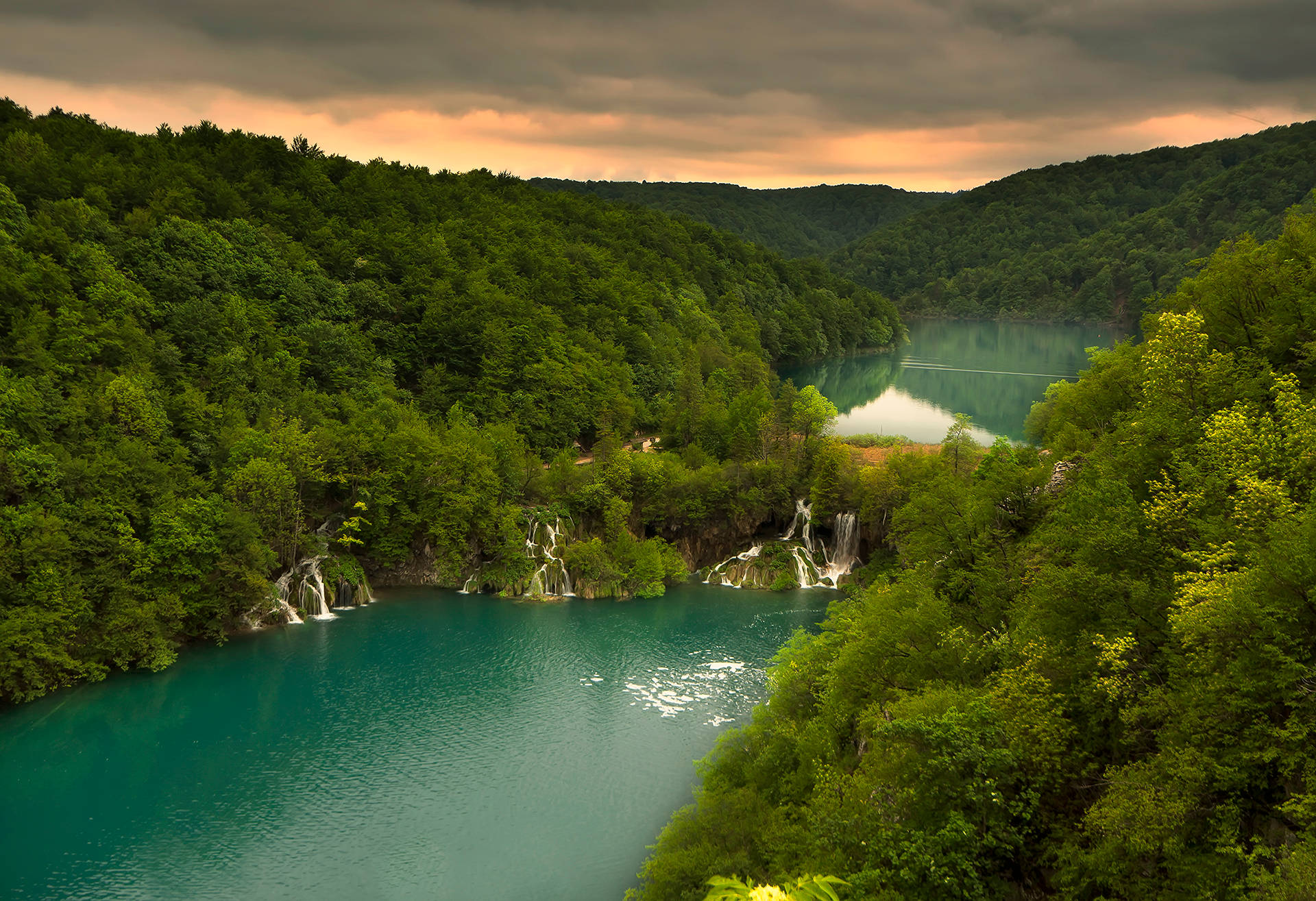 Croatia 桌布 壁紙 — 在戲劇性的多雲天空下，一個寧靜的綠松石湖 nestled 在茂密的綠色森林中，有瀑布傾瀉而下。