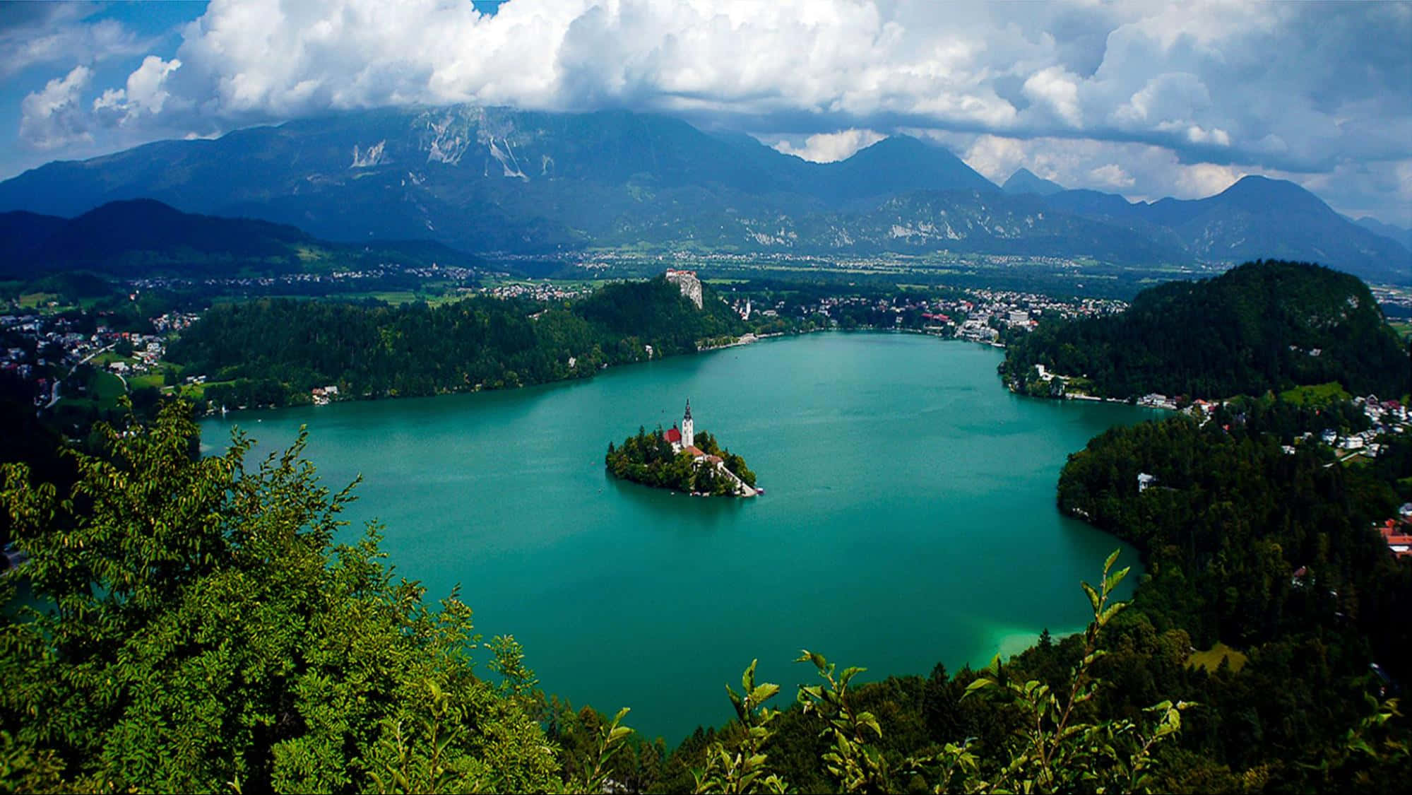Slovenia 桌布 壁紙 — 在戲劇性的多雲天空下，一個被茂密綠色森林和高聳山脈環繞的碧綠湖泊的壯麗景色。