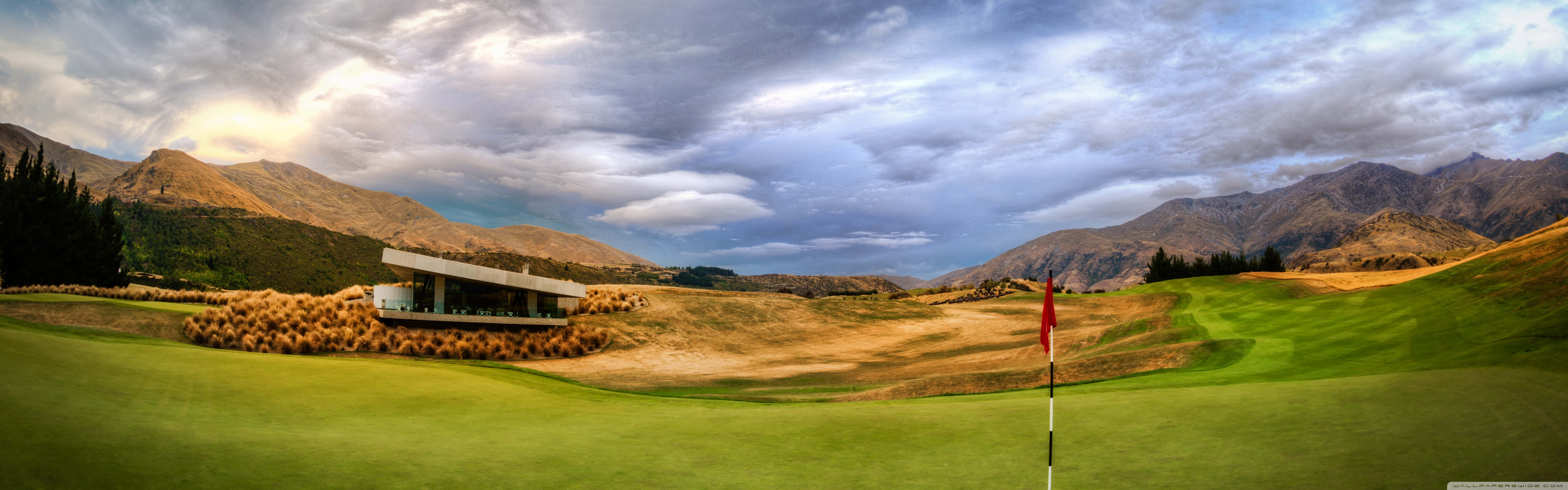 Golf 3840x1080 桌布 壁紙 — 在戲劇性的多雲天空下，一個鬱鬱蔥蔥的高爾夫球場的全景，帶有紅旗和現代會所，坐落在起伏的丘陵之間。