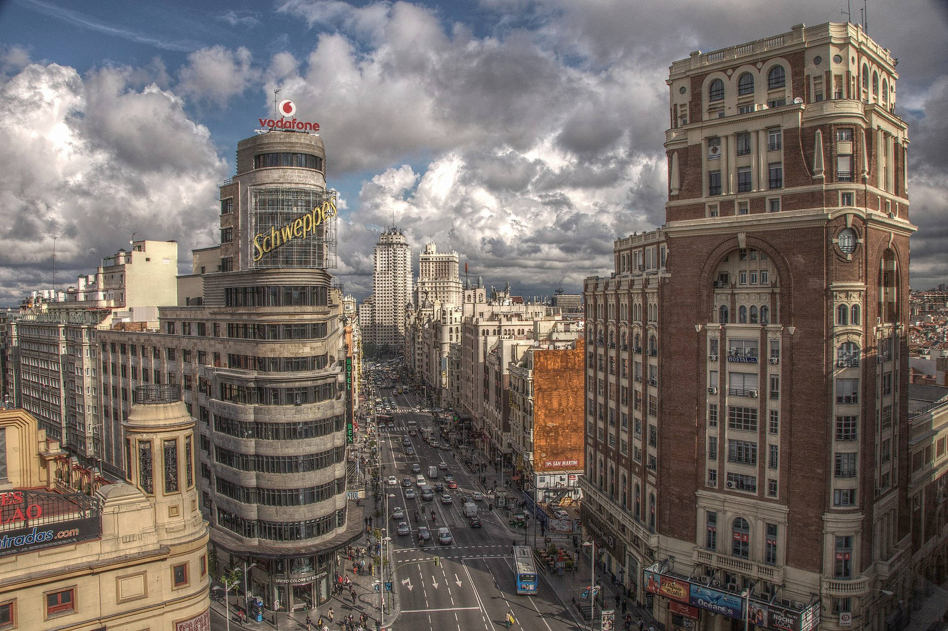 Madrid 桌布 壁紙 — 在戲劇性的多雲天空下，一條繁忙的城市街道兩旁排列著歷史和現代建築物的鳥瞰圖。