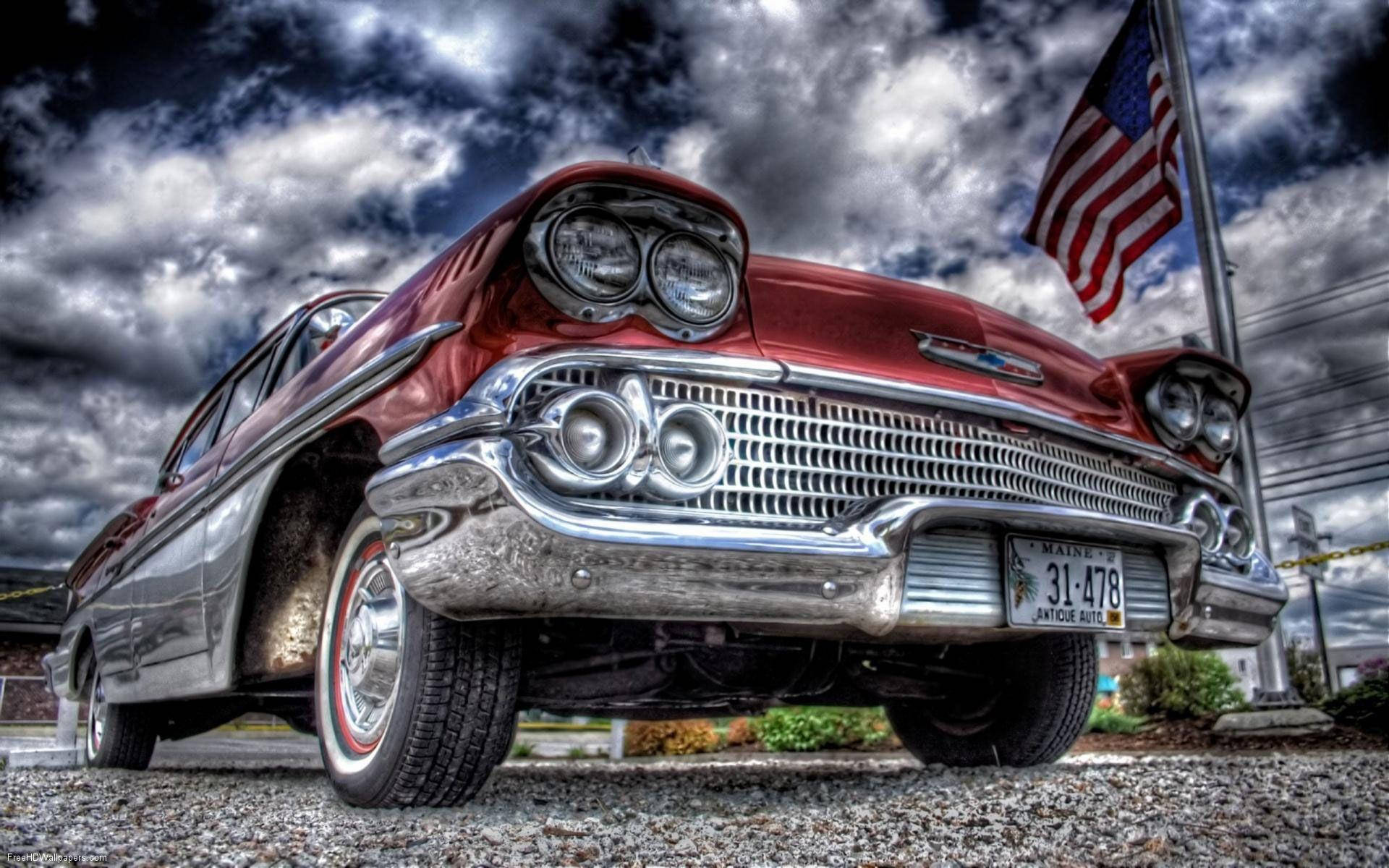 Chevrolet 桌布 壁紙 — 在戲劇性的多雲天空下，一面美國國旗飄揚，低角度拍攝一輛閃亮的紅色經典汽車，帶有鍍鉻細節。