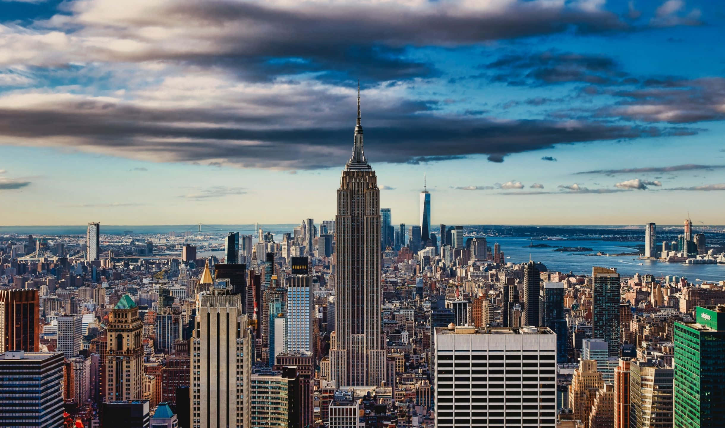 2440x1440 Empire State Building 桌布 壁紙 — 在戲劇性的多雲天空下，以帝國大廈為主導的紐約市天際線的廣闊航拍視圖。