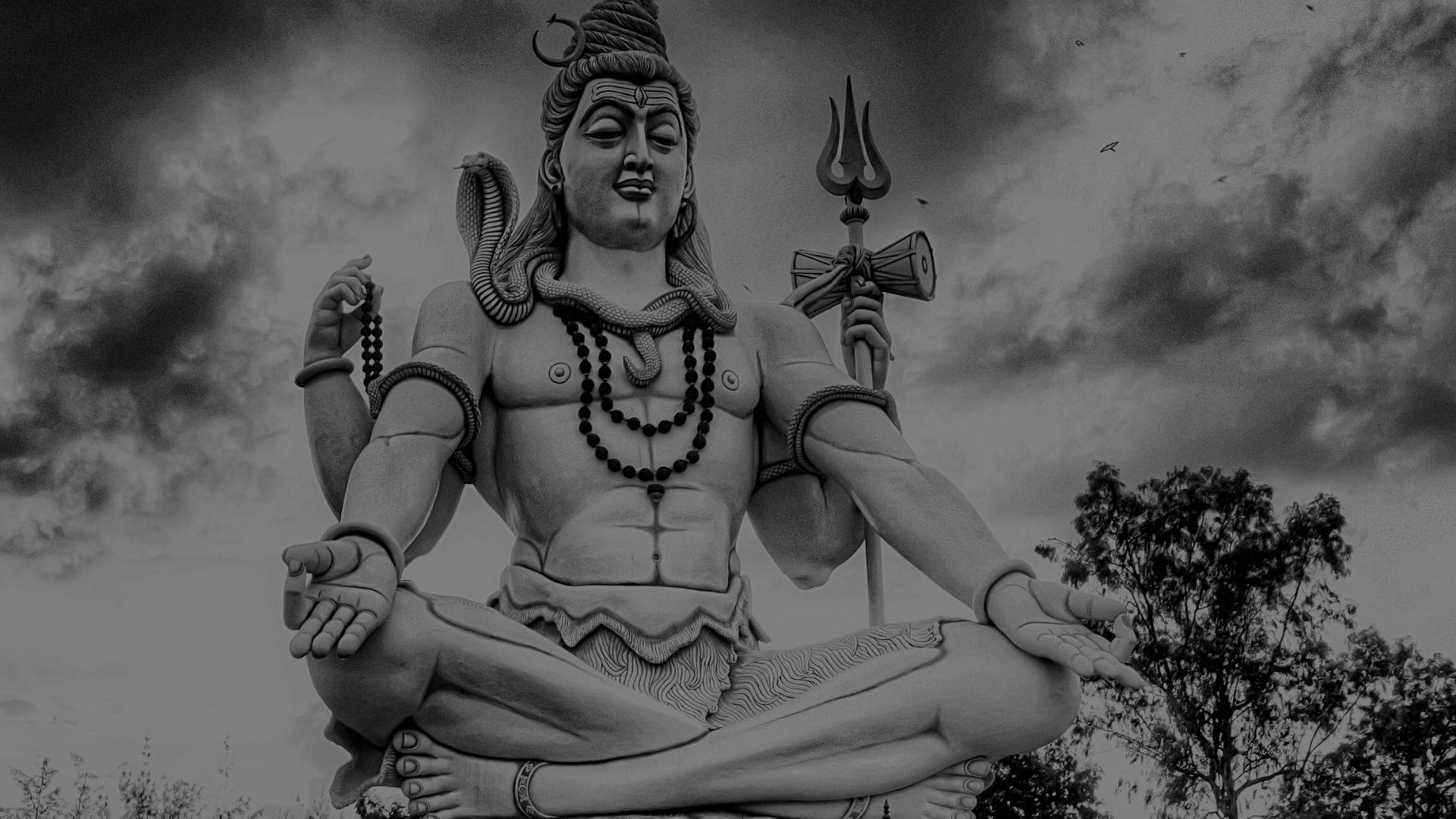 shiva dark 桌布 壁紙 — 在戲劇性的多雲天空下，濕婆神冥想姿勢手持三叉戟和念珠的紀念性黑白雕像。