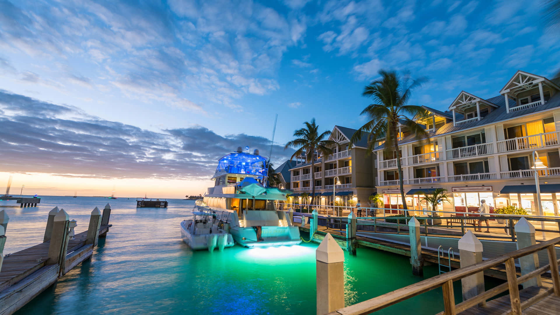 Key West 桌布 壁紙 — 在戲劇性的天空下，一艘發光的藍色船隻停泊在棕櫚樹環繞的度假村建築旁，構成充滿活力的黃昏港口場景。