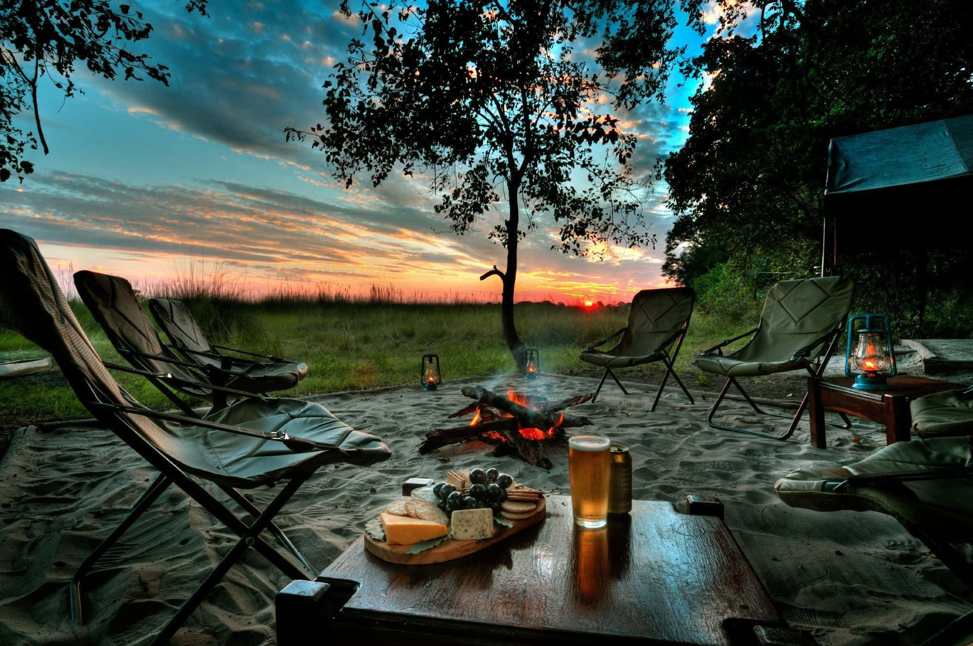 Campfire 桌布 壁紙 — 在戲劇性的天空下，營火在日落時溫暖地發光，椅子圍繞著一張放有起司、葡萄和啤酒的桌子。