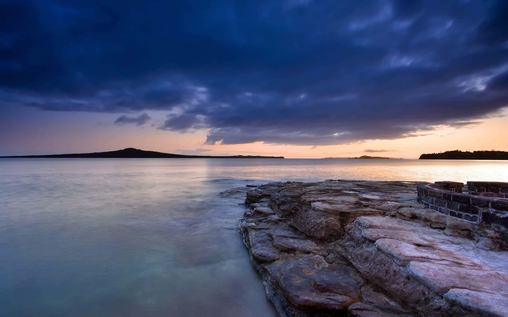 Calming Desktop 桌布 壁紙 — 在戲劇性的紫色和橙色天空下，平靜的海面流過堅固的石頭防波堤，呈現寧靜的黃昏海景。