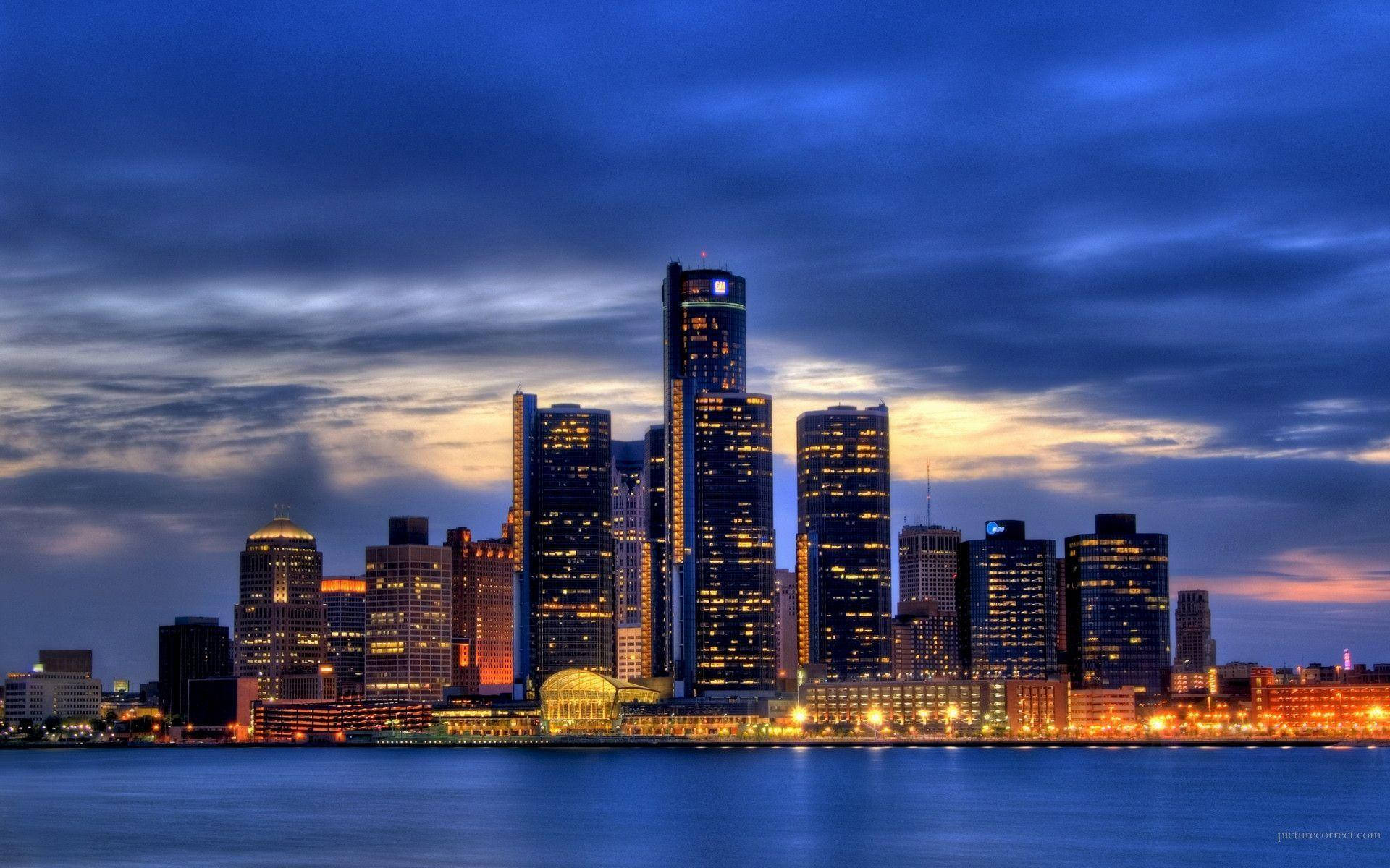 detroit 桌布 壁紙 — 在戲劇性的藍色和橙色天空下，燈火通明的摩天大樓倒映在平靜水面上，呈現出充滿活力的城市天際線。