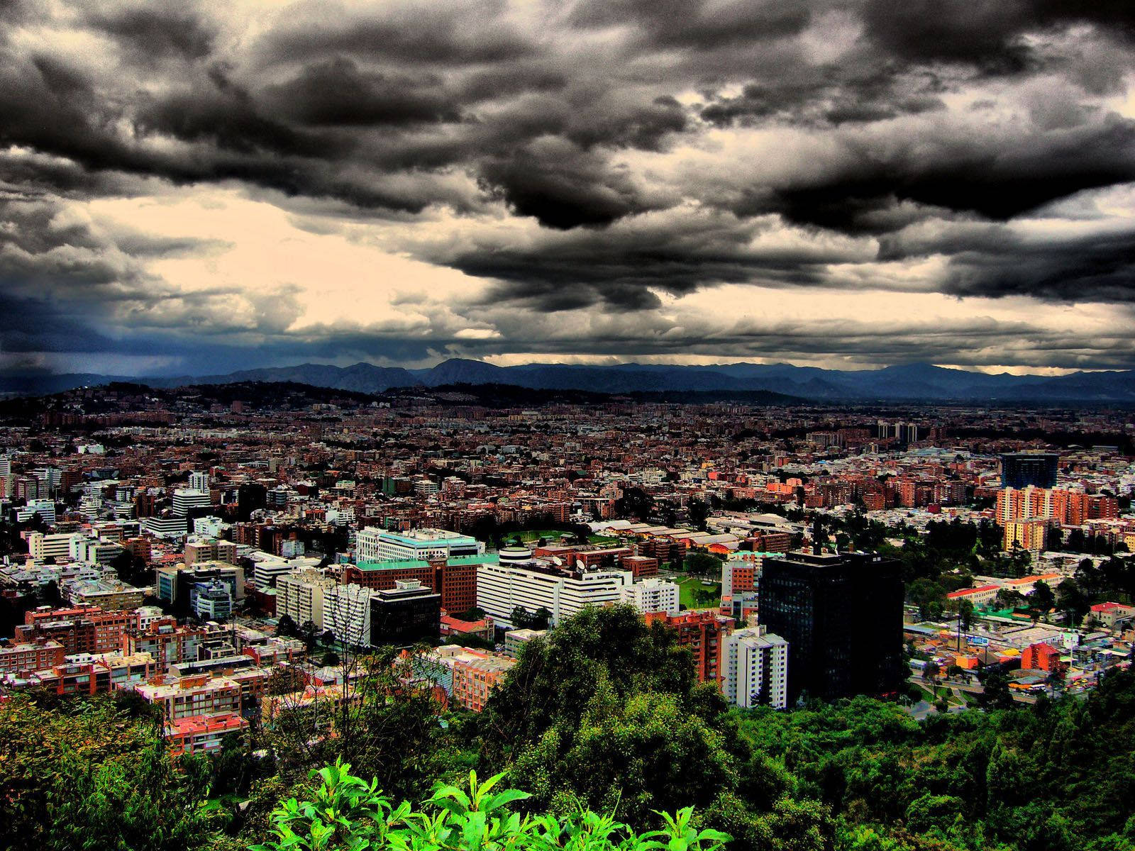 Bogota 桌布 壁紙 — 在戲劇性的風暴雲下廣闊的城市景觀，前景有茂密的綠色植物。