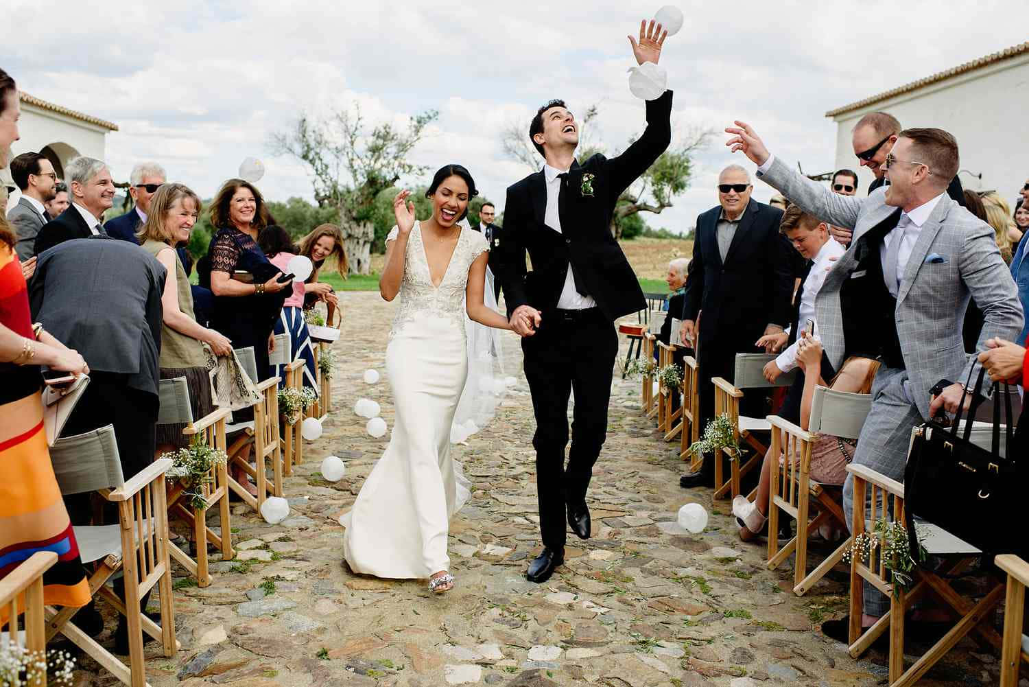 Groom And Bride 桌布 壁紙 — 在戶外婚禮上，一對快樂的新娘和新郎在飄落的白色花瓣中跳躍，賓客們沿著石徑歡呼。