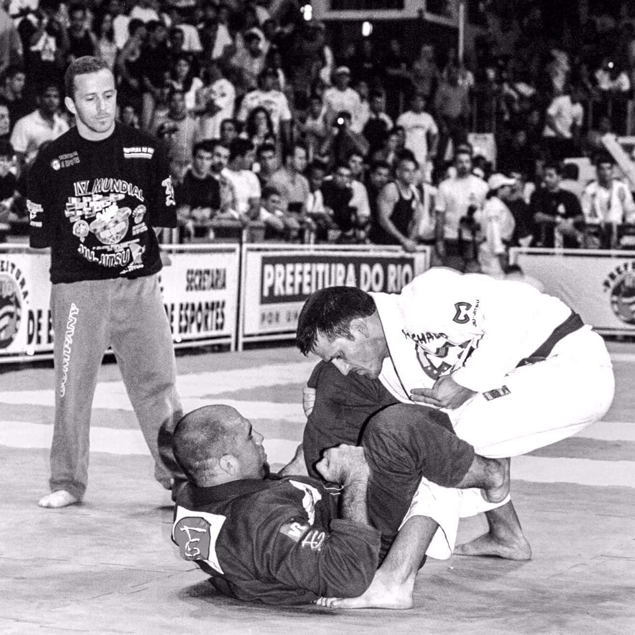 MMA 桌布 壁紙 — 在擁擠的巴西柔術比賽中，兩名選手在墊子上激烈纏鬥，裁判在一旁觀看。
