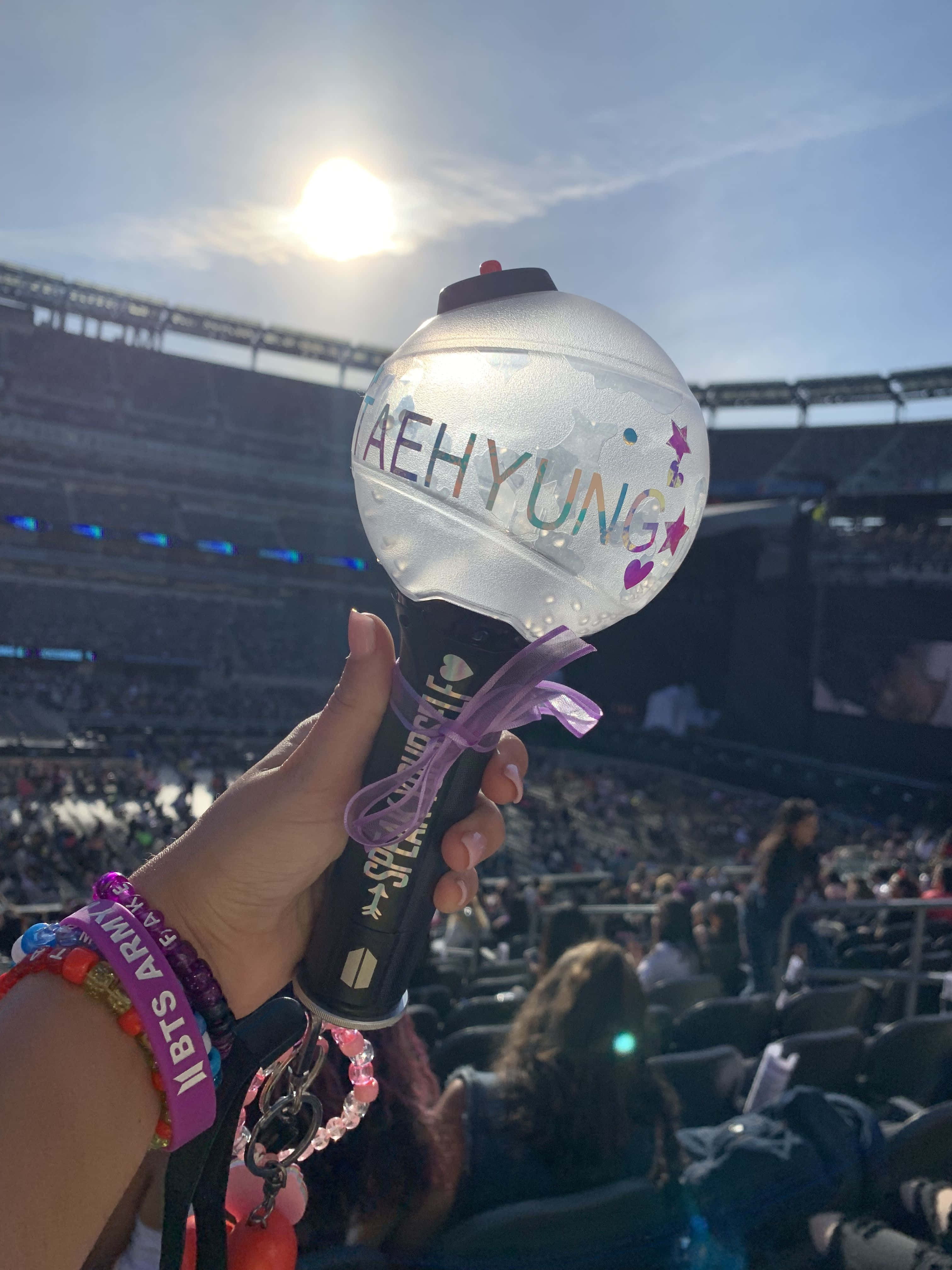 Bts Army Bomb 桌布 壁紙 — 在擁擠的體育場演唱會上，在明亮的陽光下，一隻手拿著發光的應援棒，上面寫著「AEHYUNG」。