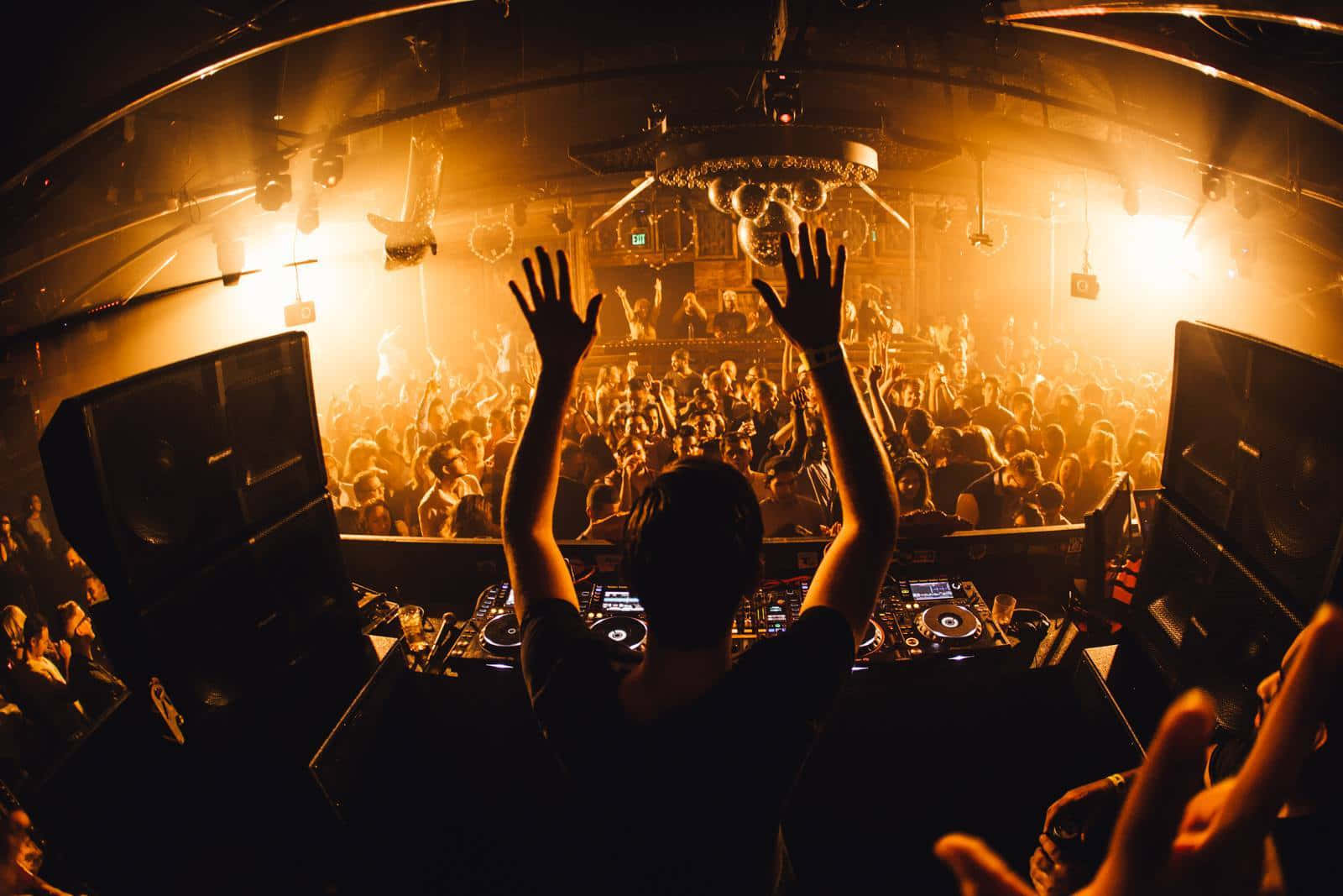 club 桌布 壁紙 — 在擠滿人群的夜店中，一位DJ舉起雙臂的剪影，金色燈光照亮下方跳舞的人群。