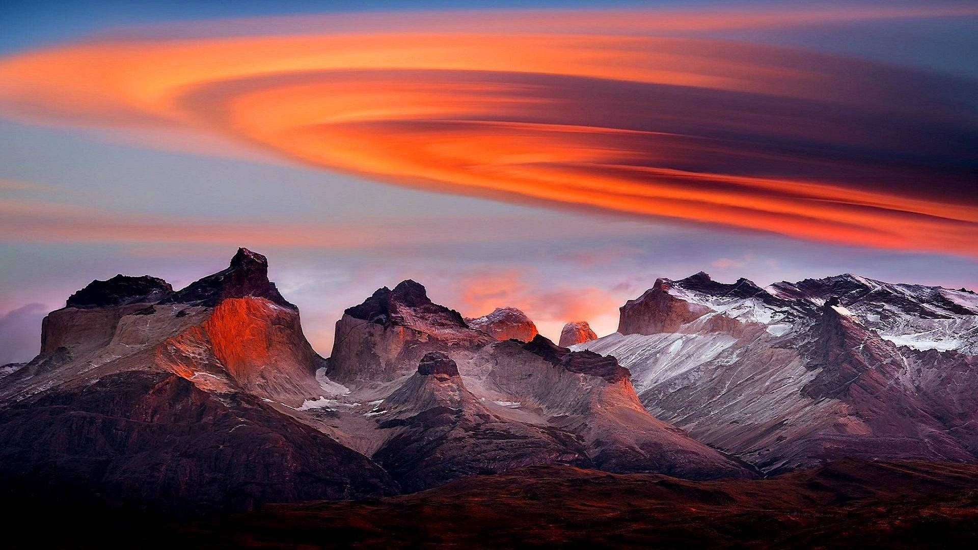 Patagonia 桌布 壁紙 — 在旋轉的橙色和紫色天空下，一條戲劇性的山脈沐浴在熾熱的日落色調中。