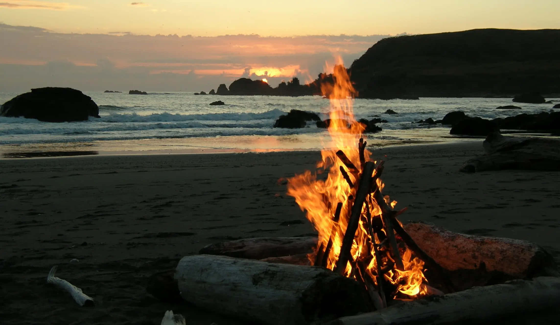 Bonfire 桌布 壁紙 — 在日落時分，黑暗的沙灘上熊熊燃燒的營火，背景中波浪拍打岩石和山丘。