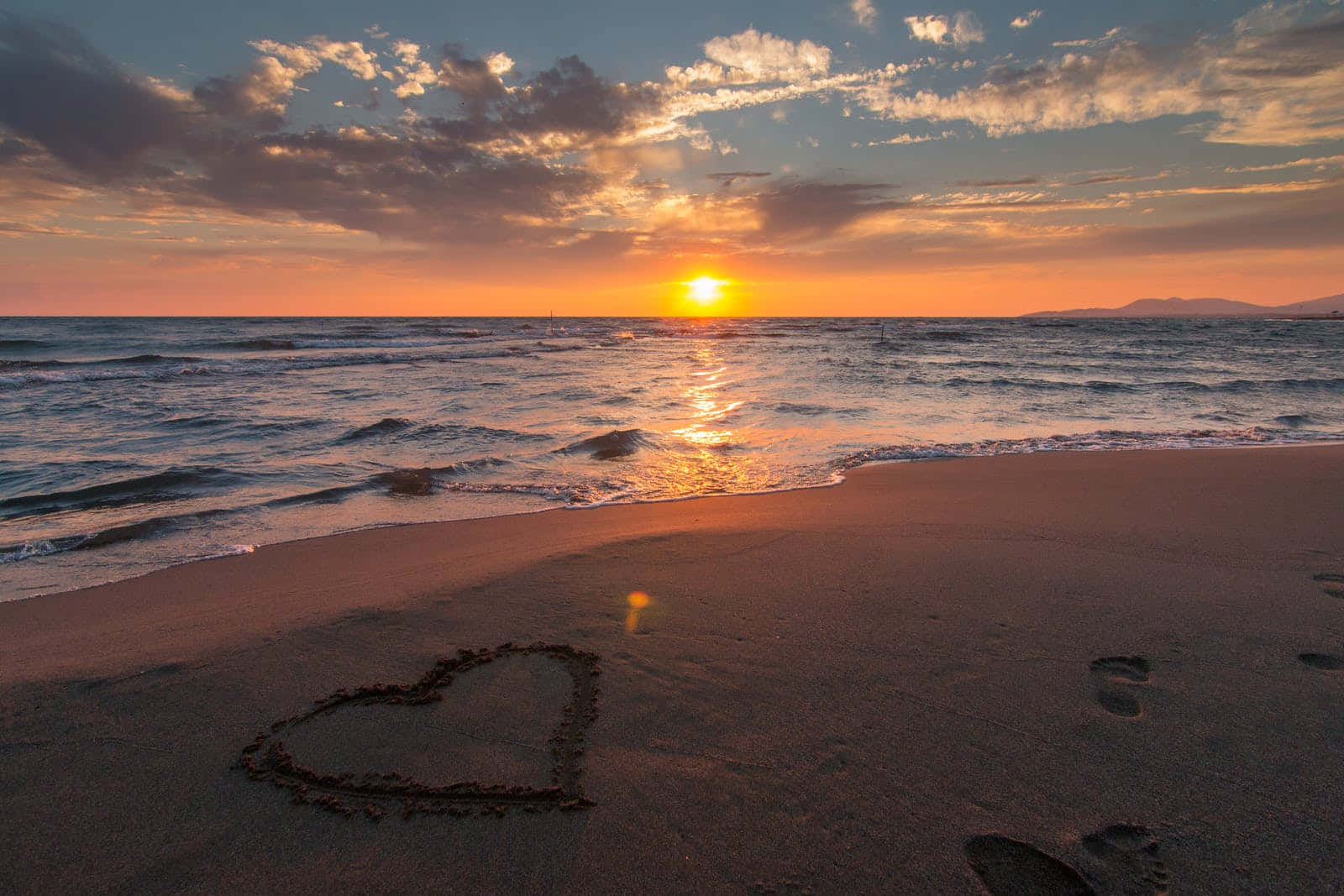 Cute Sunset 桌布 壁紙 — 在日落時分於濕沙上畫的心形，金色光線反射在海浪和遠處的山巒上。
