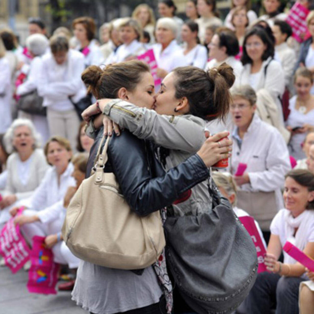 Kissing 桌布 壁紙 — 在日間抗議活動中，兩名女性在舉著粉紅色標語牌的人群面前熱情擁抱和親吻。