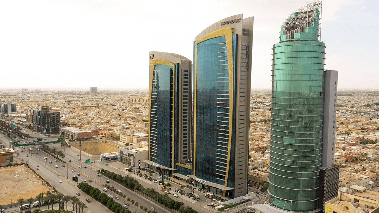 Riyadh 桌布 壁紙 — 在明亮日光下，現代玻璃摩天大樓帶金色點綴，在廣闊沙漠城市景觀上空升起的鳥瞰圖。