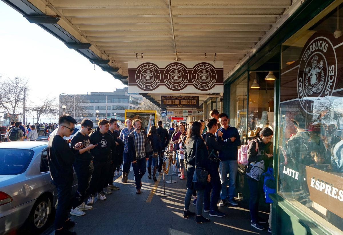 Pike Place Market 桌布 壁紙 — 在明亮的日光下，一輛停好的車附近，各種各樣的人在星巴克外面排著長隊。