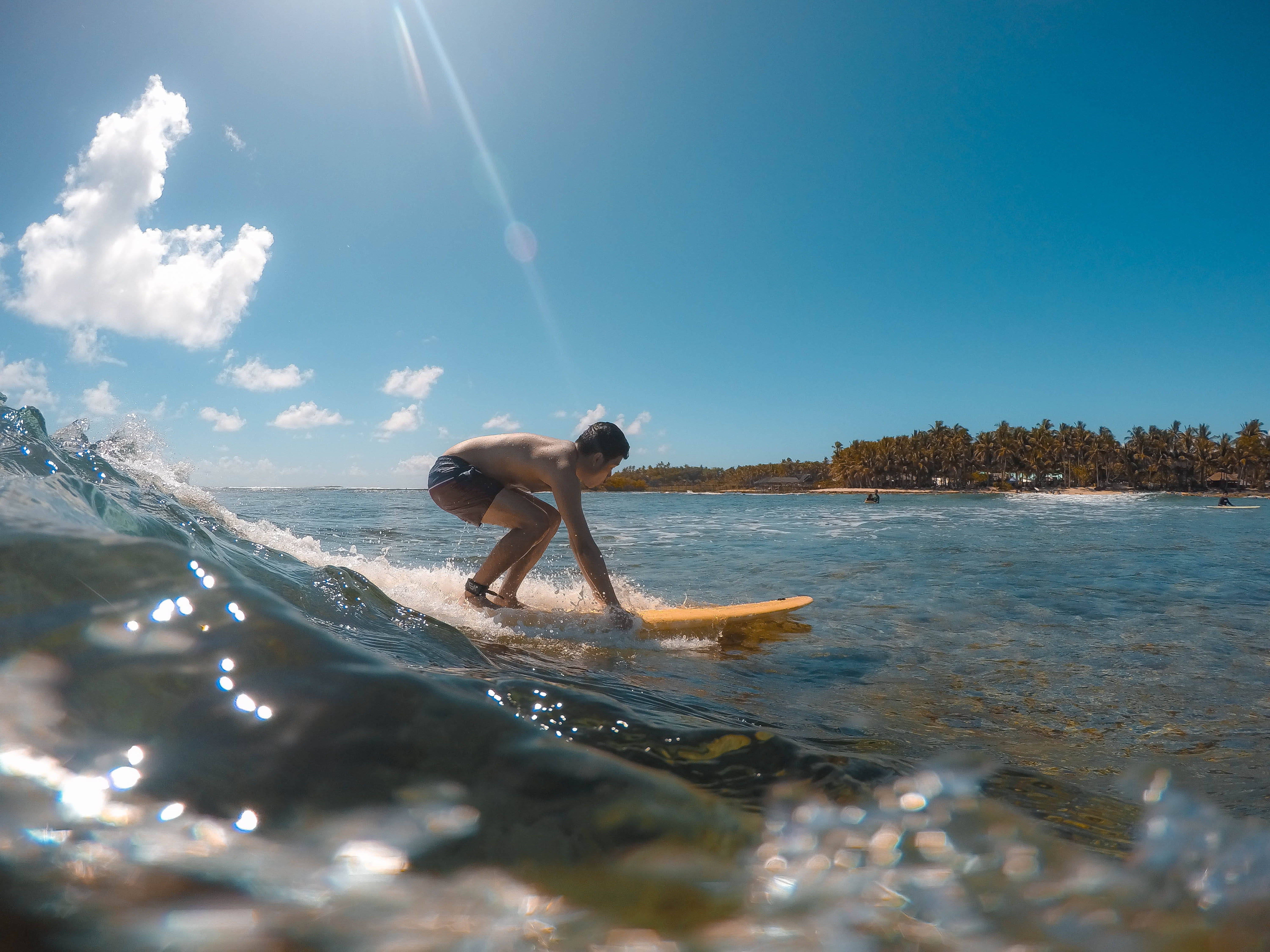Siargao 桌布 壁紙 — 在明亮的藍天下，一名衝浪者騎著小浪，背景是熱帶島嶼和棕櫚樹。