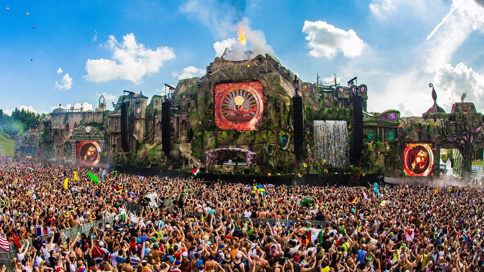 Tomorrowland 桌布 壁紙 — 在明亮的藍天下，一大群人在一個巨大的奇幻主題音樂節舞台上歡呼，舞台上有發光的螢幕和瀑布。