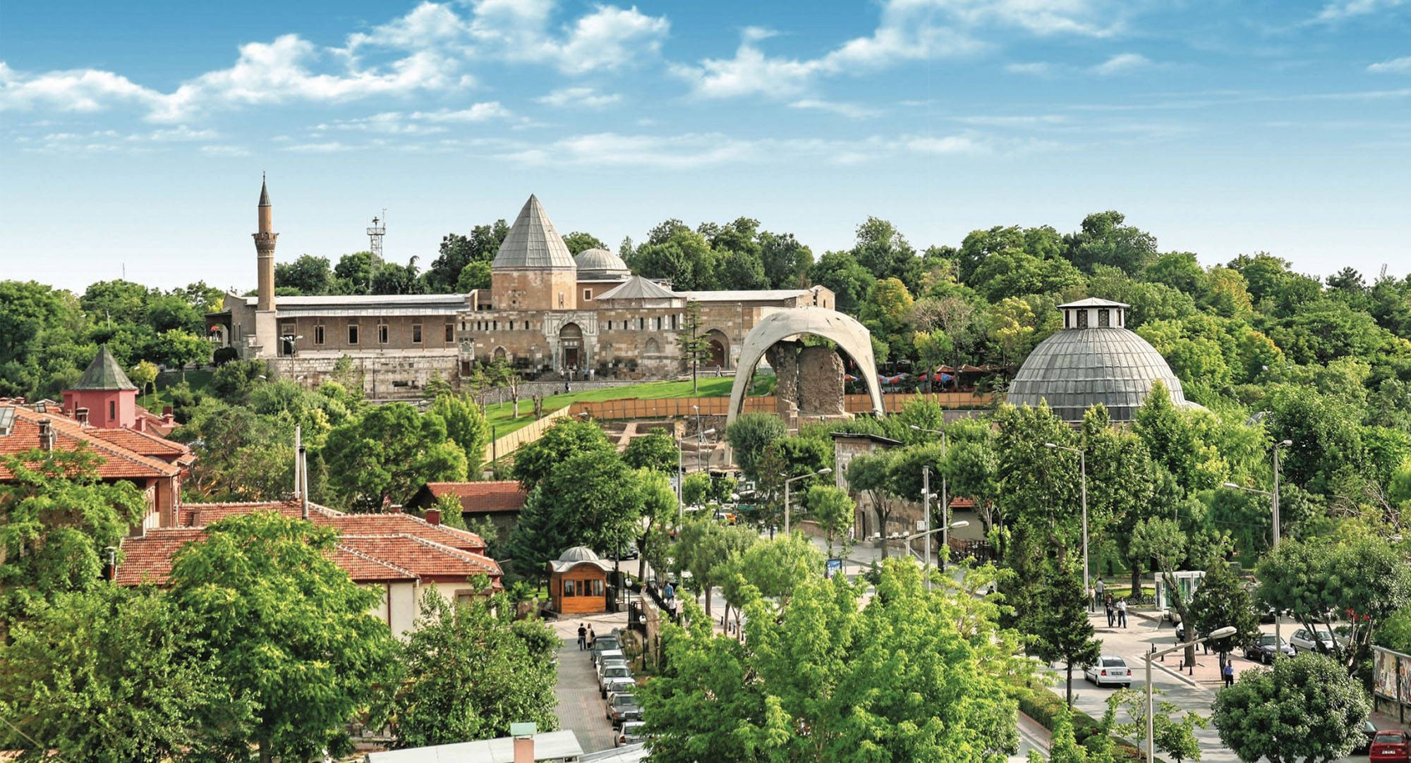 Konya 桌布 壁紙 — 在明亮的藍天下，歷史悠久的石造建築帶有圓頂和宣禮塔，隱藏在茂密的綠樹之中，呈現全景視角。