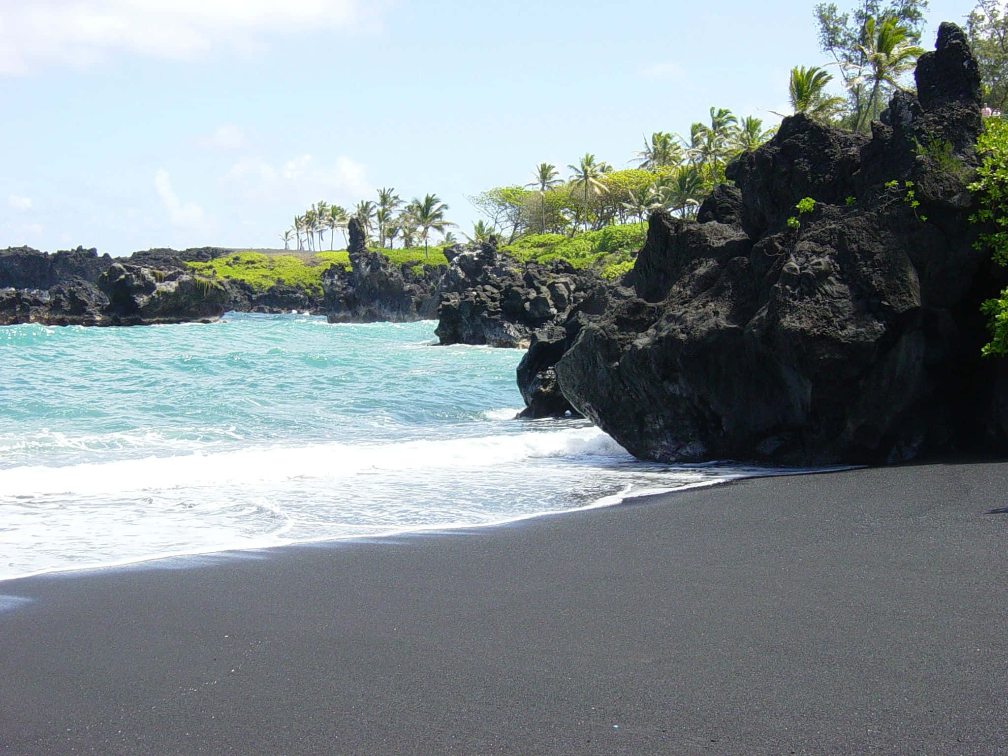 Black Sand Beach 桌布 壁紙 — 在明亮的藍天下，碧綠色的海浪拍打著黑色沙灘，旁邊是崎嶇的火山岩和茂密的棕櫚樹。