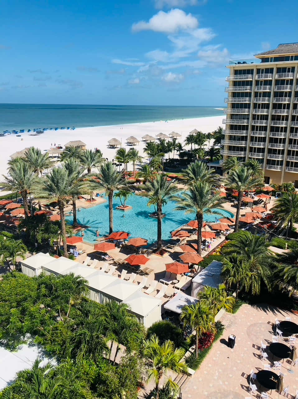 Marco Island 桌布 壁紙 — 在明亮的藍天下，豪華海灘度假村的鳥瞰圖，設有藍綠色游泳池、棕櫚樹和白色沙灘。