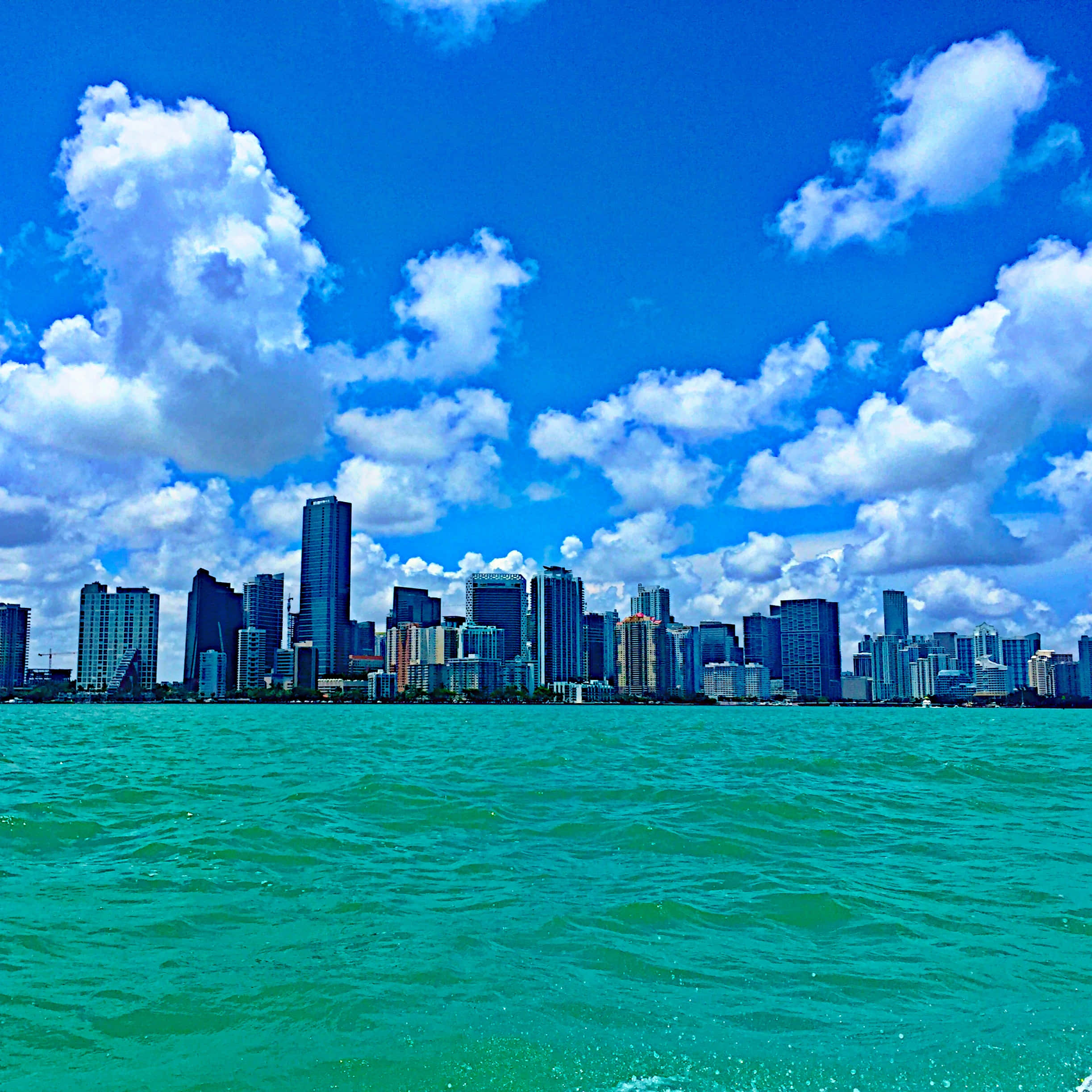 Miami 桌布 壁紙 — 在明亮的藍天下，點綴著蓬鬆的白雲，充滿活力的藍綠色水波紋勾勒出現代城市的天際線。