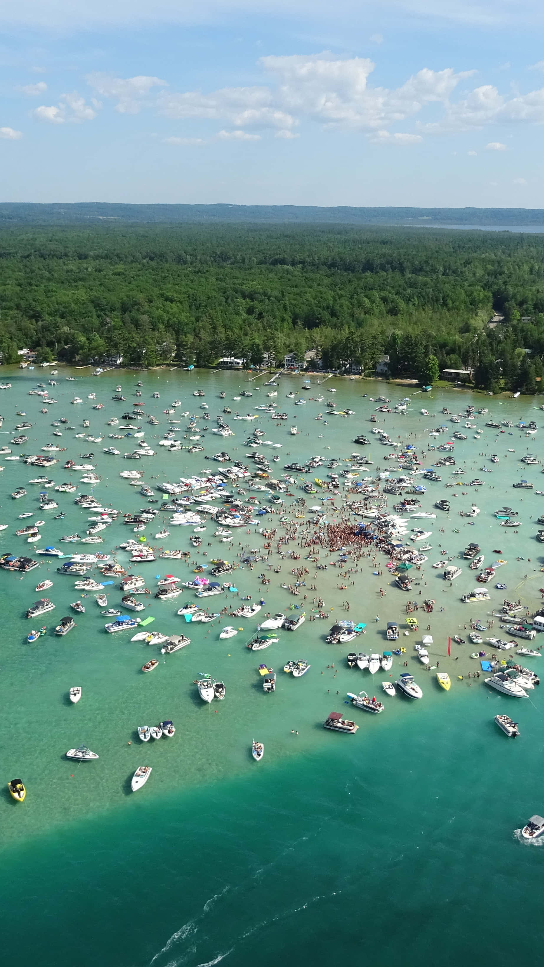 Torch Lake 桌布 壁紙 — 在明亮的藍天和零星雲朵下，擠滿船隻和游泳者的綠松石湖的鳥瞰圖。