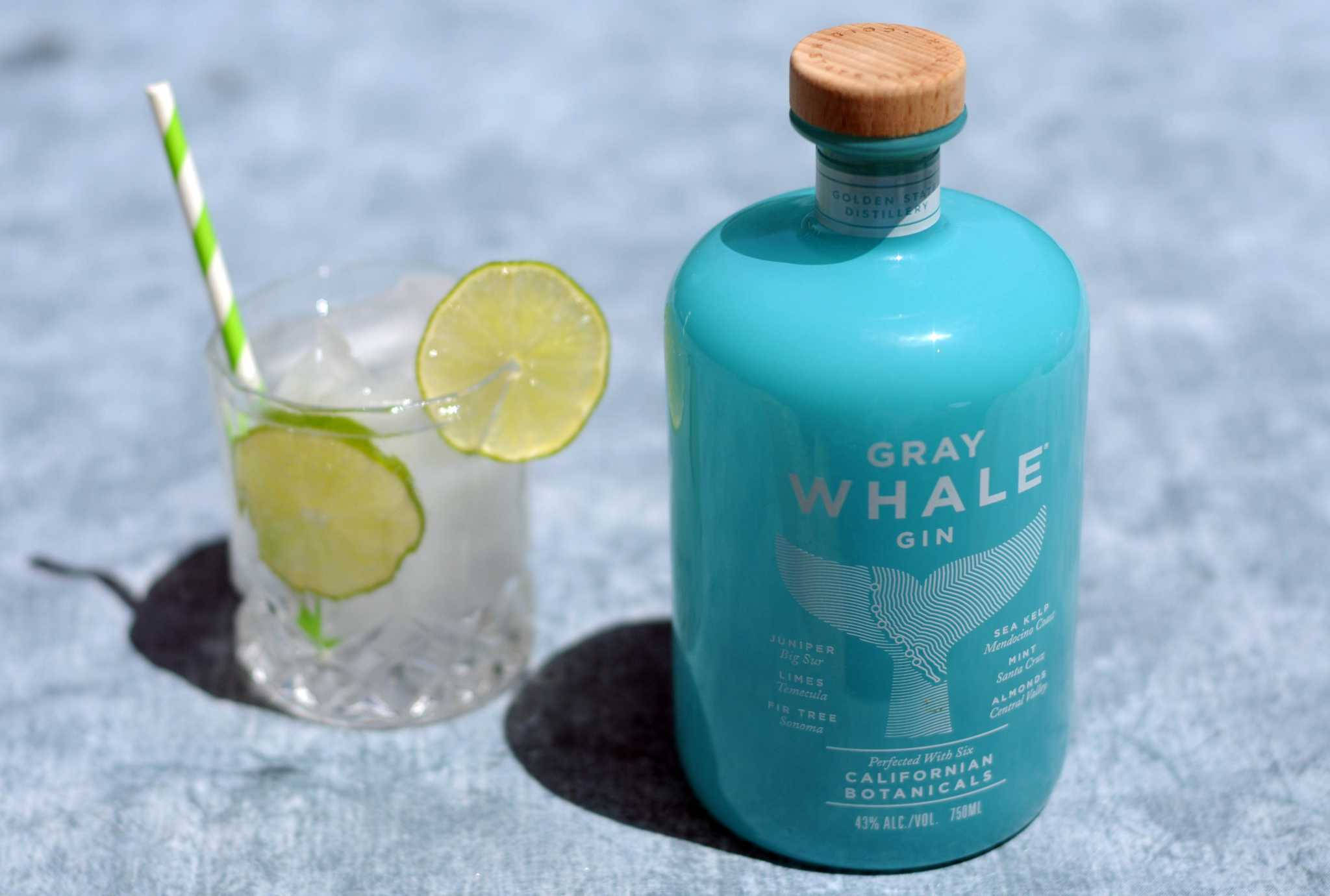 alcohol 桌布 壁紙 — 在明亮的陽光下，一個藍綠色的Gray Whale Gin酒瓶和一個以萊姆裝飾的琴酒雞尾酒放在有紋理的藍色表面上。