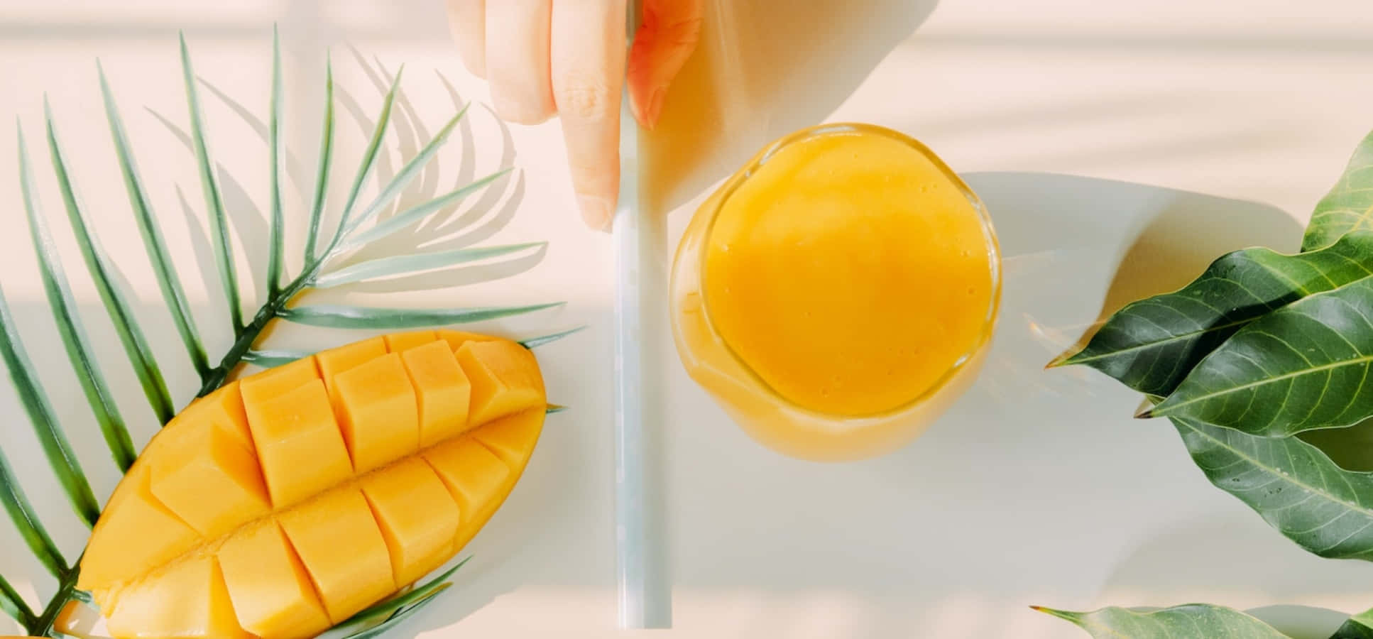 Mango 桌布 壁紙 — 在明亮的陽光下，一隻手將充滿活力的芒果汁倒入玻璃杯中，旁邊放著切片芒果和熱帶葉子。
