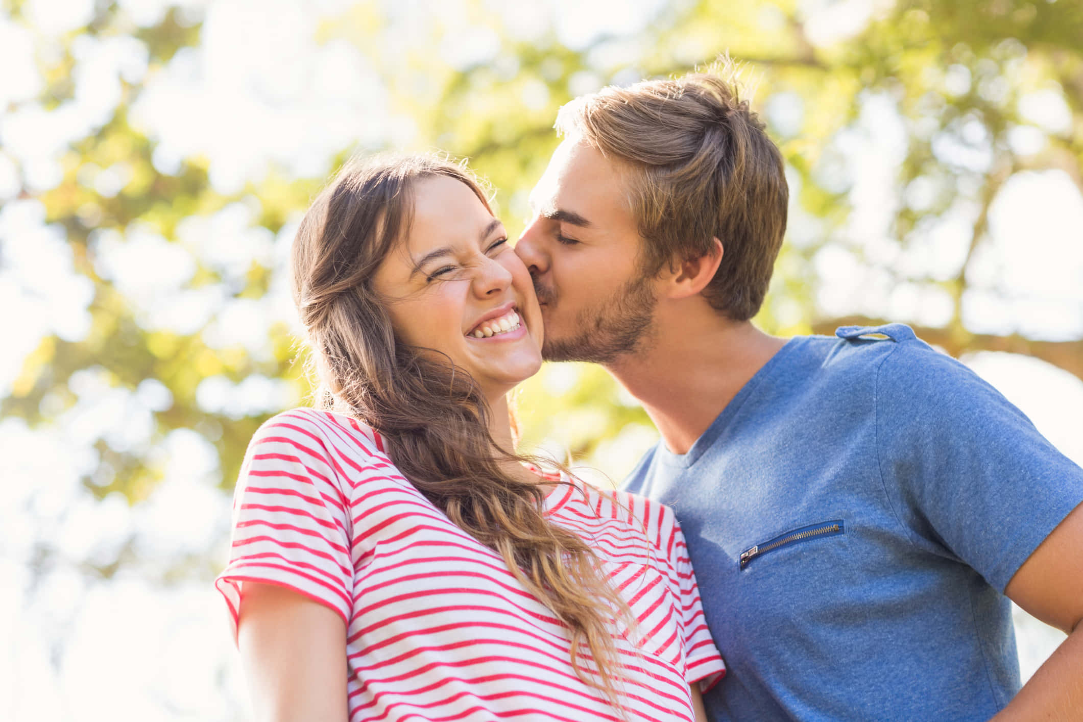 cheek kiss 桌布 壁紙 — 在明亮的陽光樹蔭下，一名男子親吻一名大笑女子的臉頰，背景是模糊的綠色樹葉。