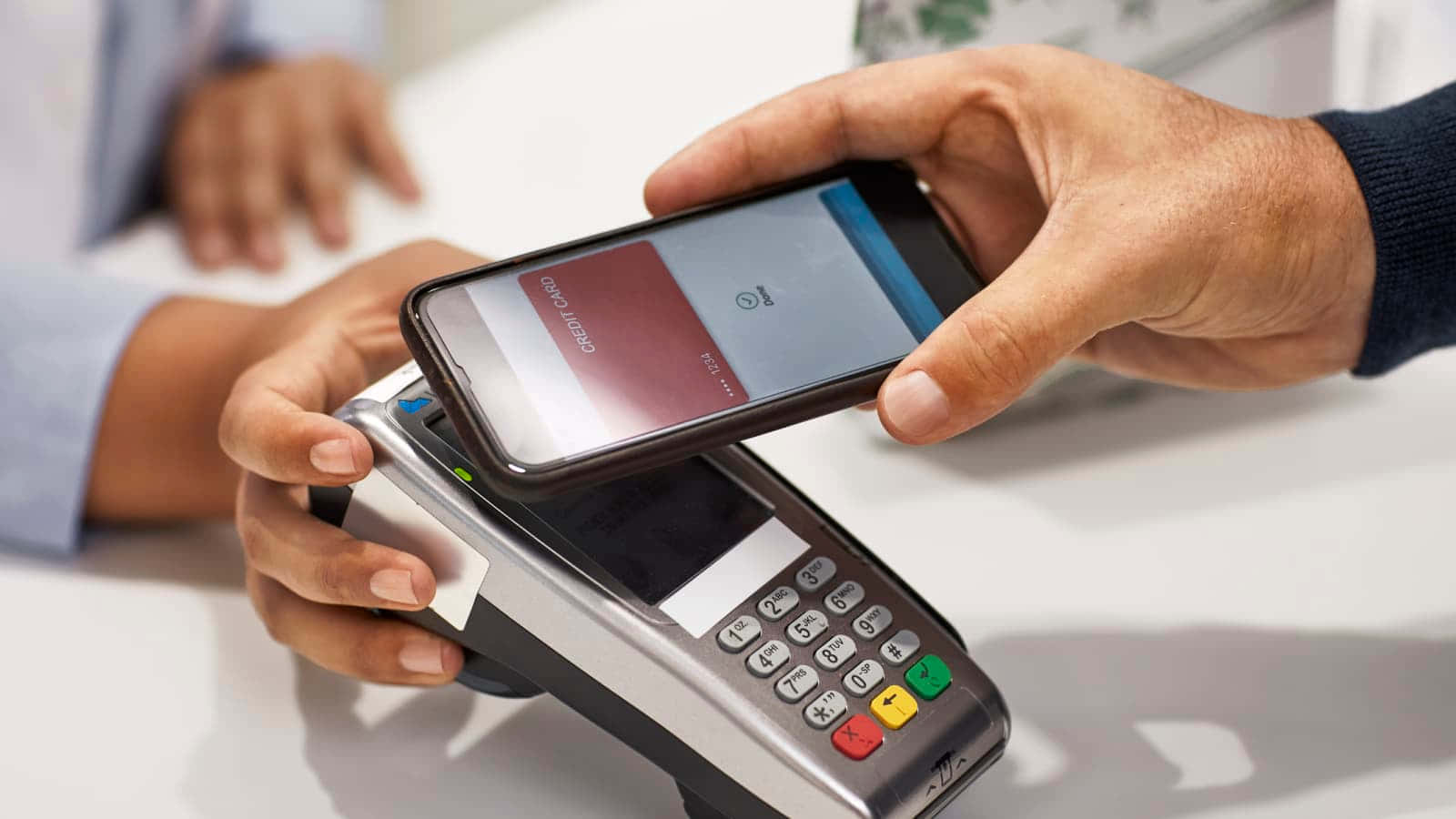 Smartphone 桌布 壁紙 — 在明亮的零售環境中，一個人將智慧型手機輕觸信用卡讀卡機進行非接觸式付款。