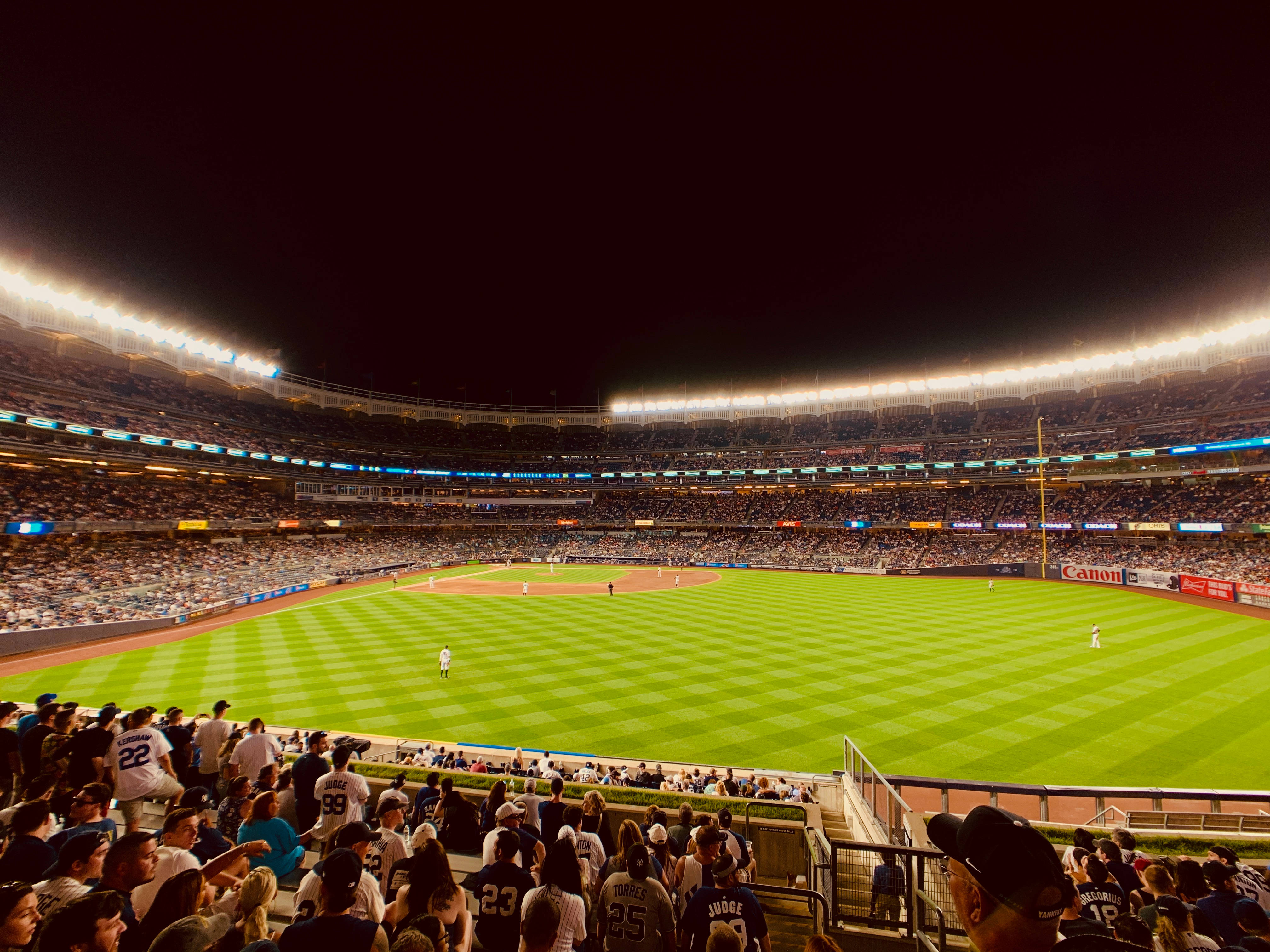 MLB 桌布 壁紙 — 在明亮的體育場燈光下進行的充滿活力的夜間棒球比賽，前景有球迷，精心條紋的綠色場地上有球員。