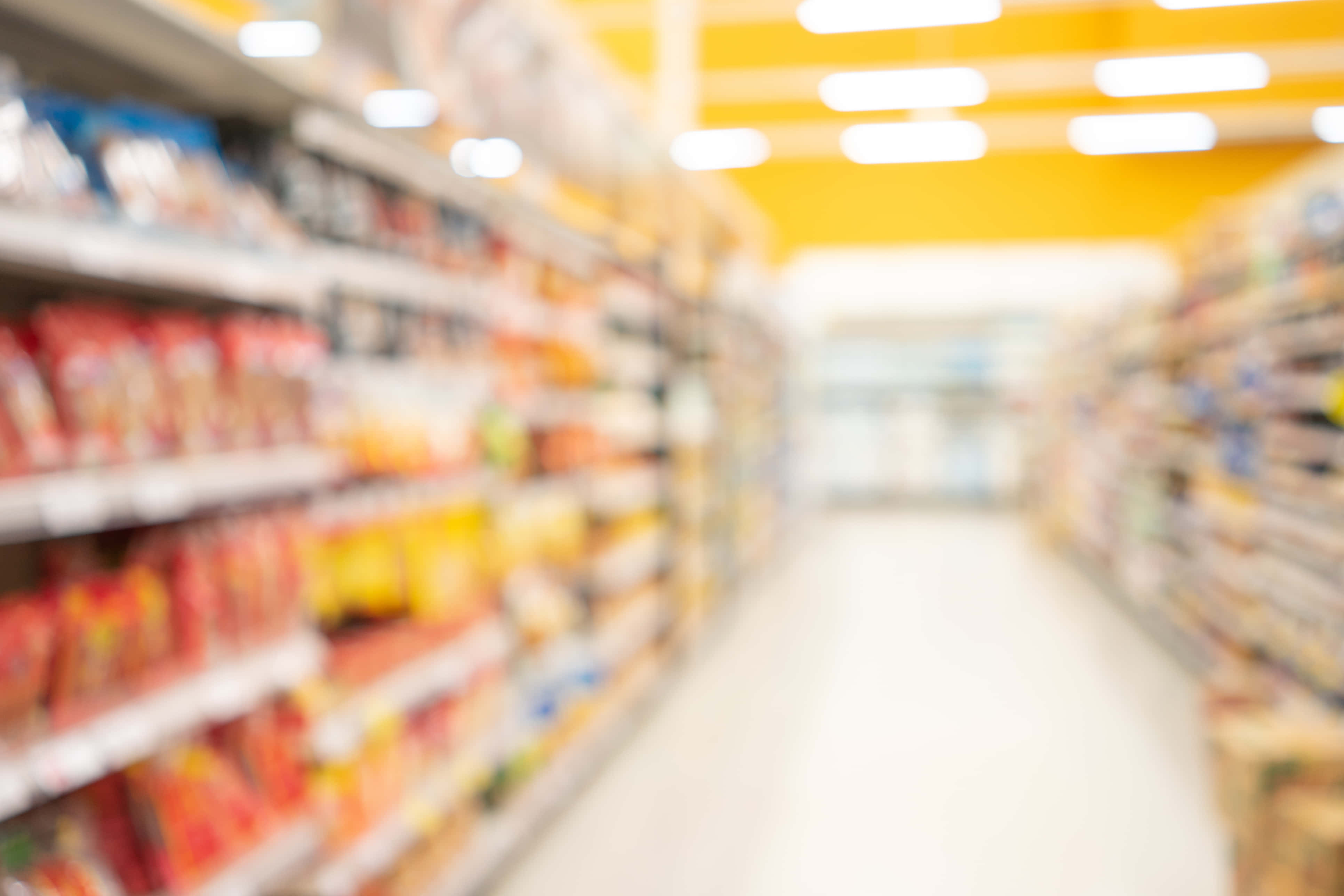 Grocery Store 桌布 壁紙 — 在明亮的黃色天花板燈光下，模糊的超級市場通道，擺滿色彩繽紛的產品貨架。