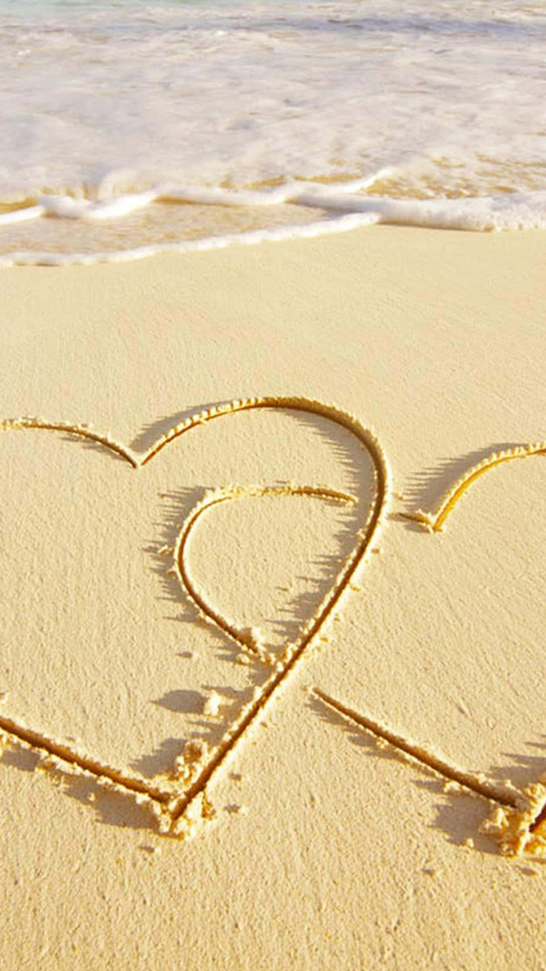 Beach Love 桌布 壁紙 — 在明亮陽光下，溫柔的海浪拍打岸邊，濕沙上畫著一顆心。