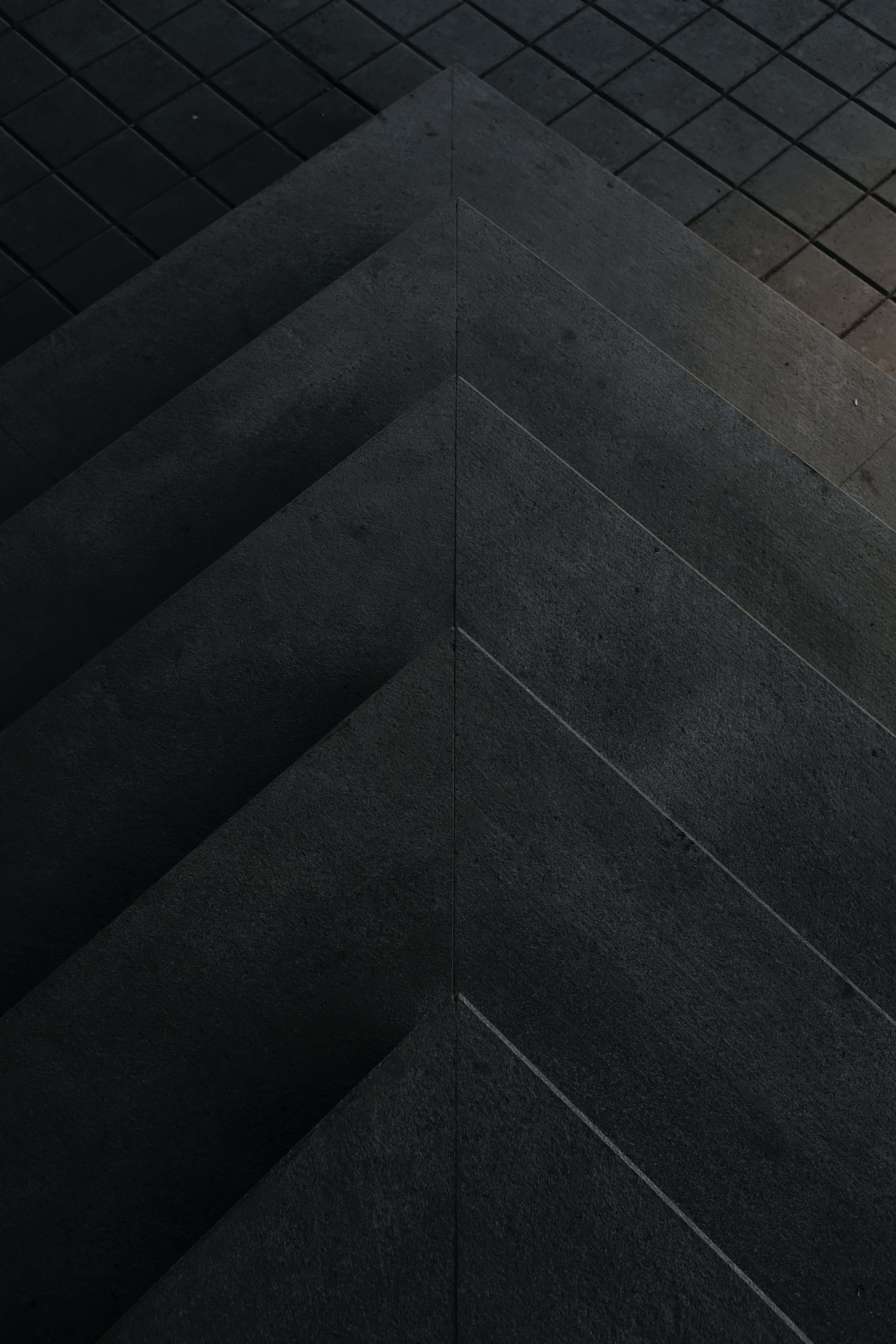 Geometric Pattern 桌布 壁紙 — 在昏暗光線下，深灰色混凝土階梯在瓷磚鋪砌的路面上形成銳利的人字形圖案的近距離特寫。