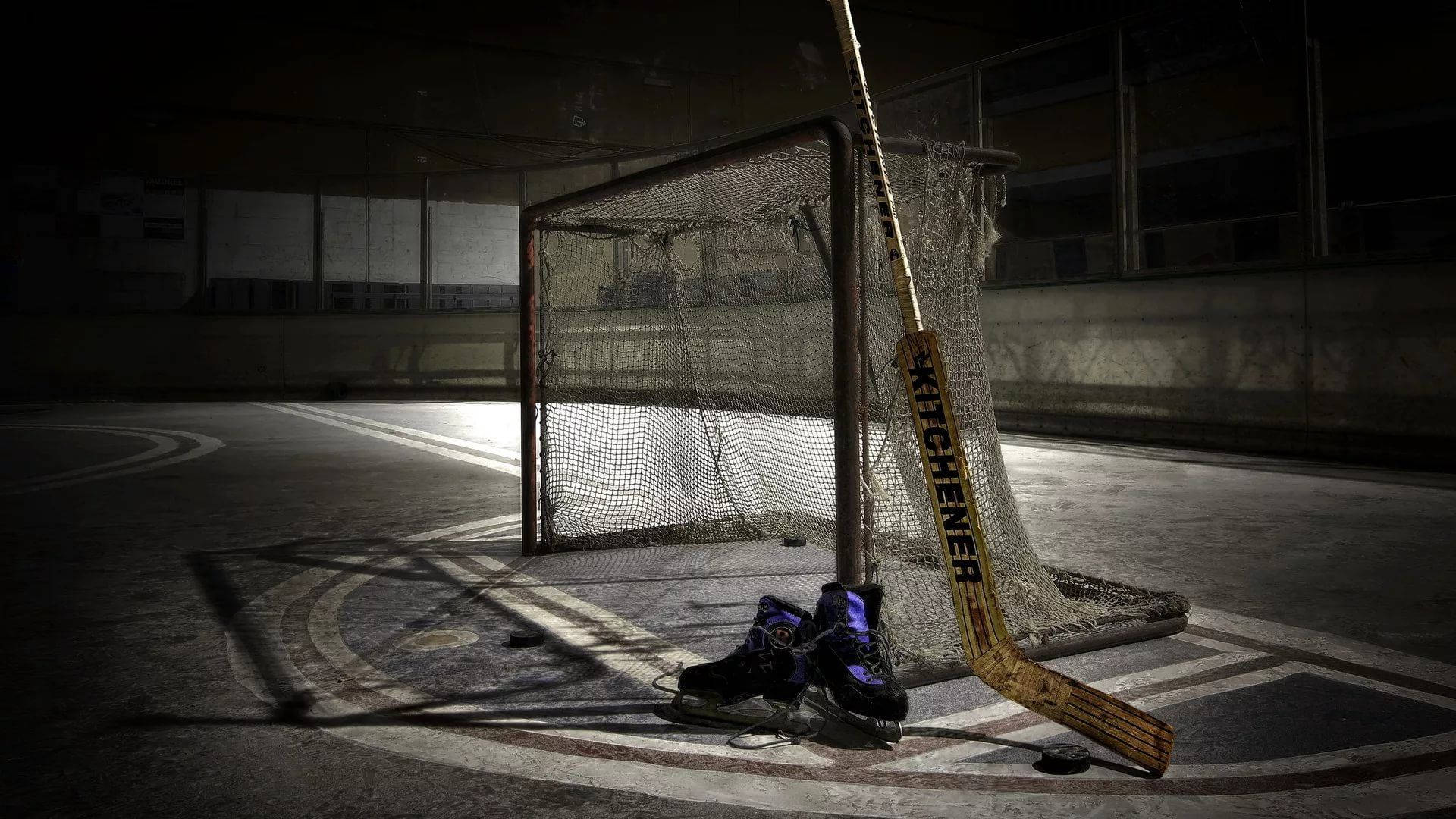 hockey 桌布 壁紙 — 在昏暗燈光下，一支孤獨的曲棍球棍和溜冰鞋放在一個廢棄溜冰場的破舊球網旁。