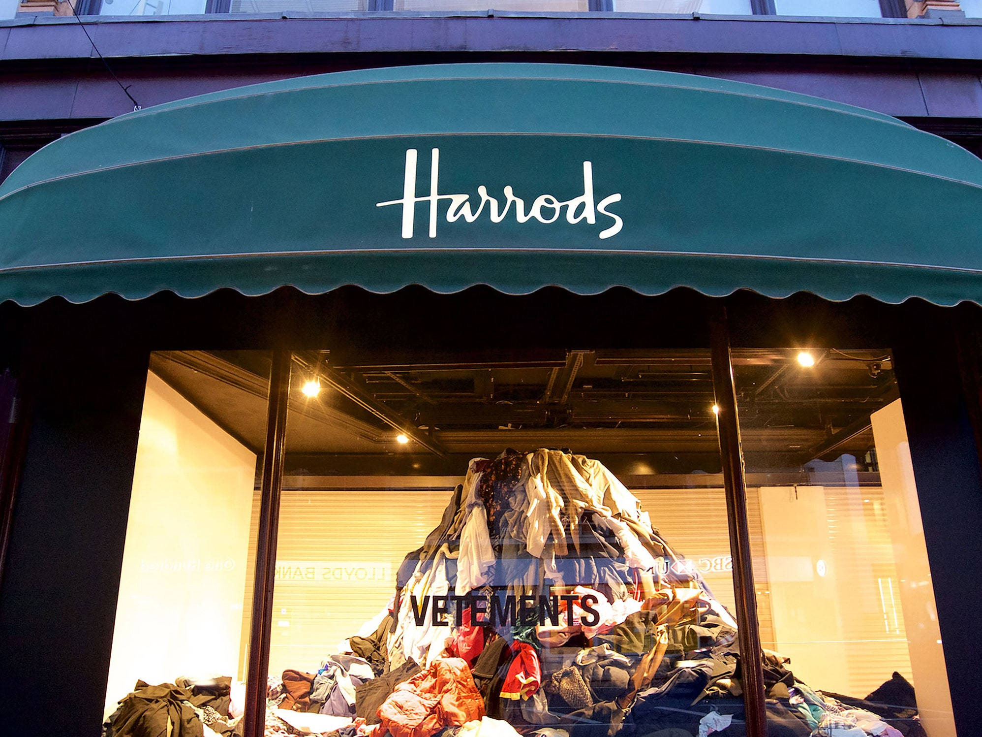 Vetements 桌布 壁紙 — 在昏暗的商店櫥窗中，一堆色彩繽紛的衣物在綠色Harrods遮陽篷下形成如山般的展示。