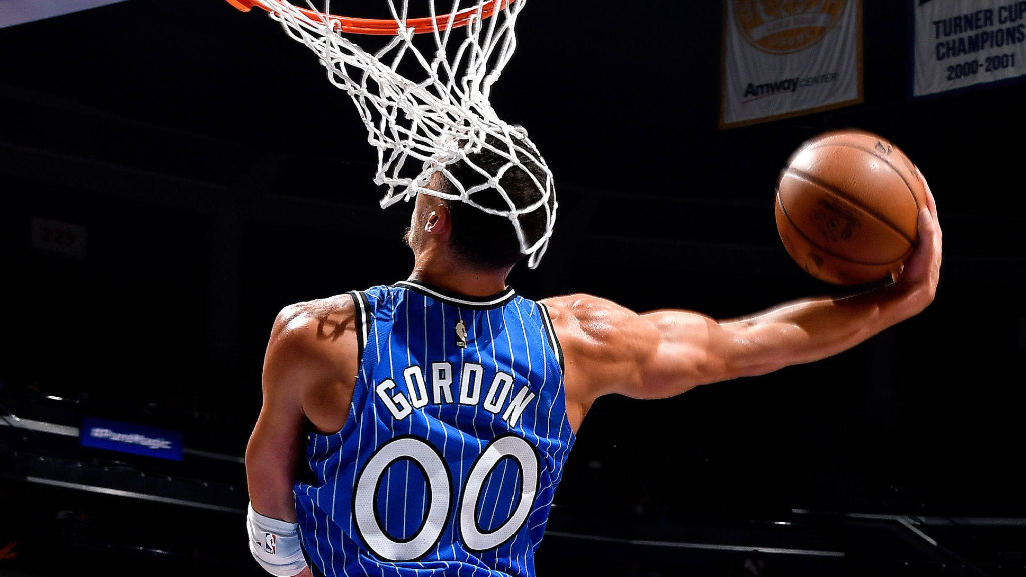 aaron gordon 桌布 壁紙 — 在昏暗的場館中，一名身穿藍色細條紋球衣的籃球運動員扣籃，籃網在頭頂搖擺。
