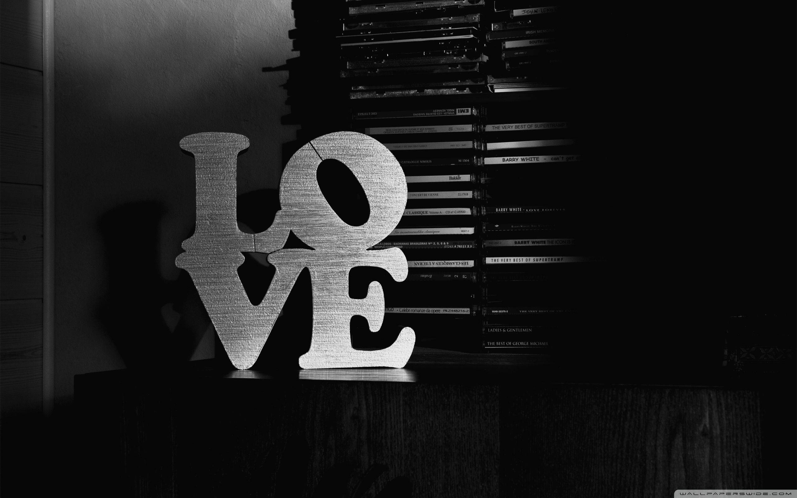 黑色愛情 桌布 壁紙 — 在昏暗的房間裡，一個單色的「LOVE」標誌在木架上柔和地發光，旁邊是一疊高聳的DVD。