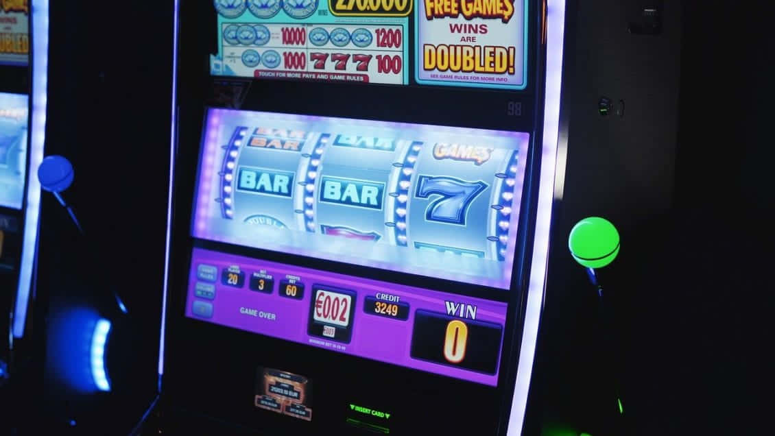 game 桌布 壁紙 — 在昏暗的賭場環境中，一部明亮照明的老虎機散發著霓虹燈光和勝利符號。
