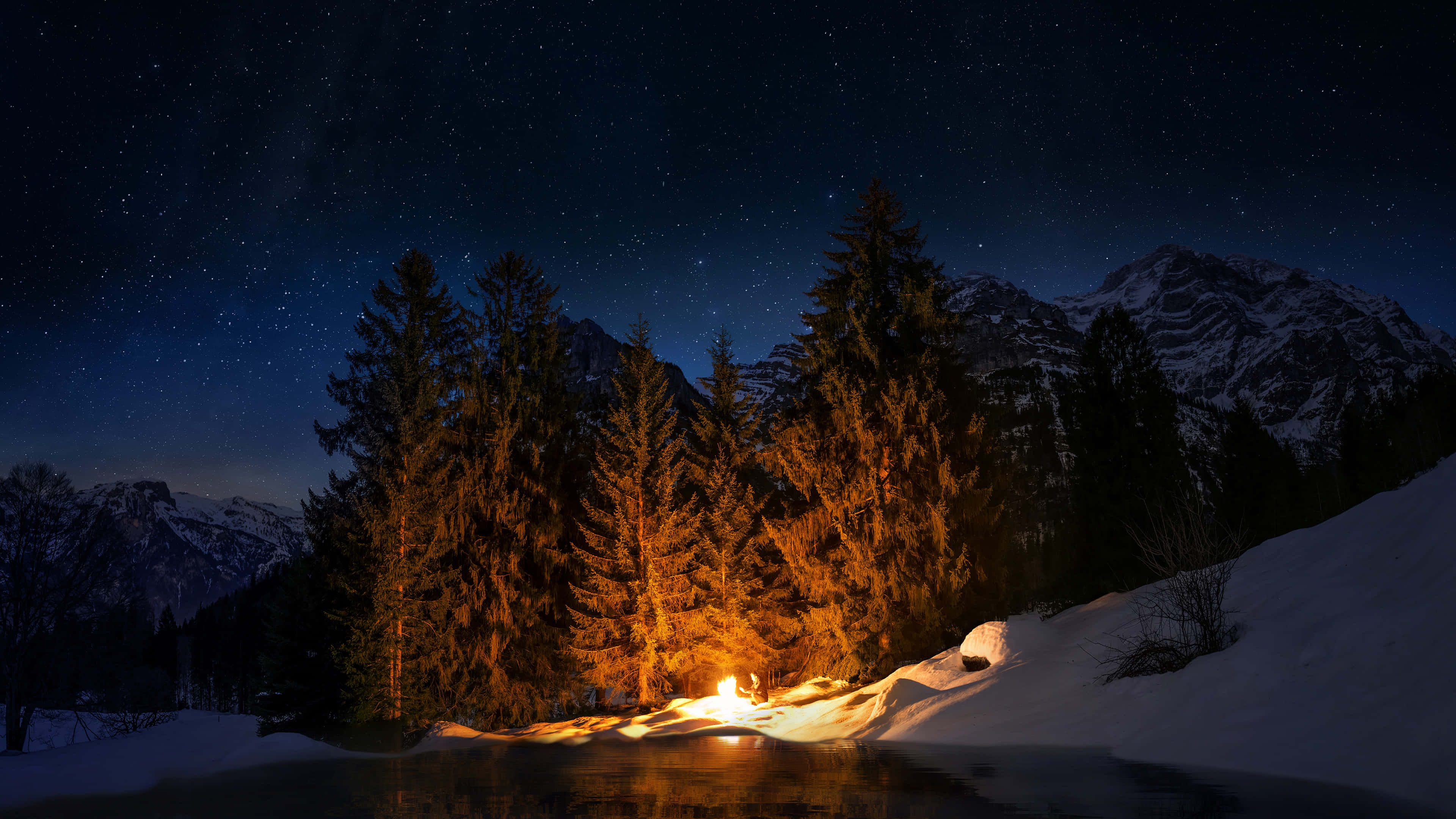 Camping 桌布 壁紙 — 在星空下，一個孤獨的營火在雪湖旁溫暖地燃燒，周圍有松樹和遠山。