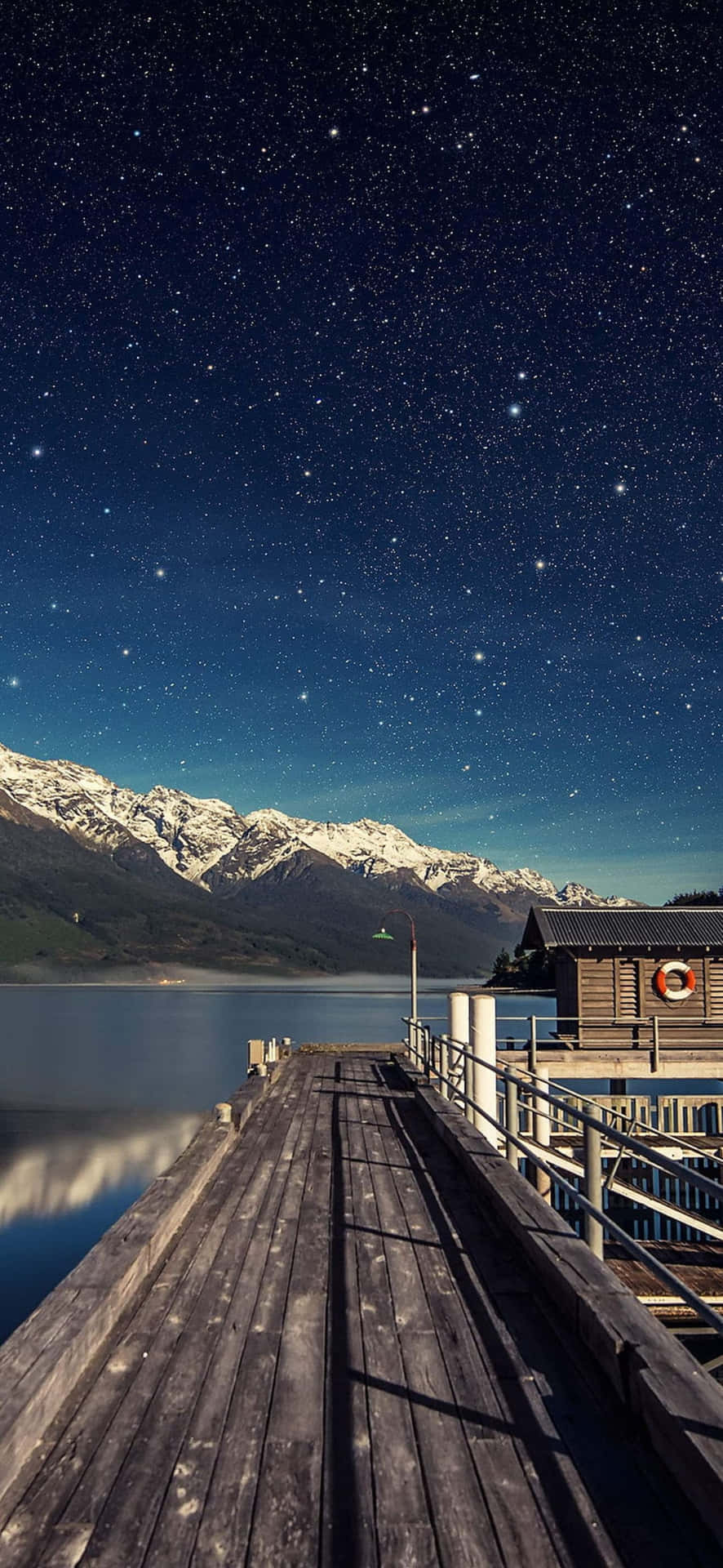 夜晚的木製碼頭，上方有星星，平靜的水面反射著雪山。 — Google pixel 壁紙